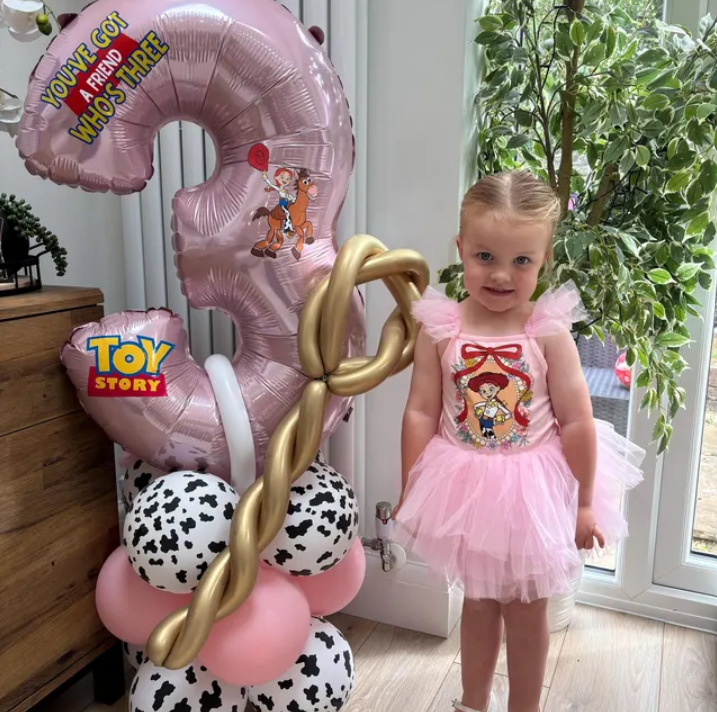 Young girl in pink dress with a Toy Story print, standing next to a balloon arrangement featuring a large pink number 3, a Toy Story balloon, gold and white balloons, and cow print balloons. Disney Party celebrations, birthday party dress, Jessy