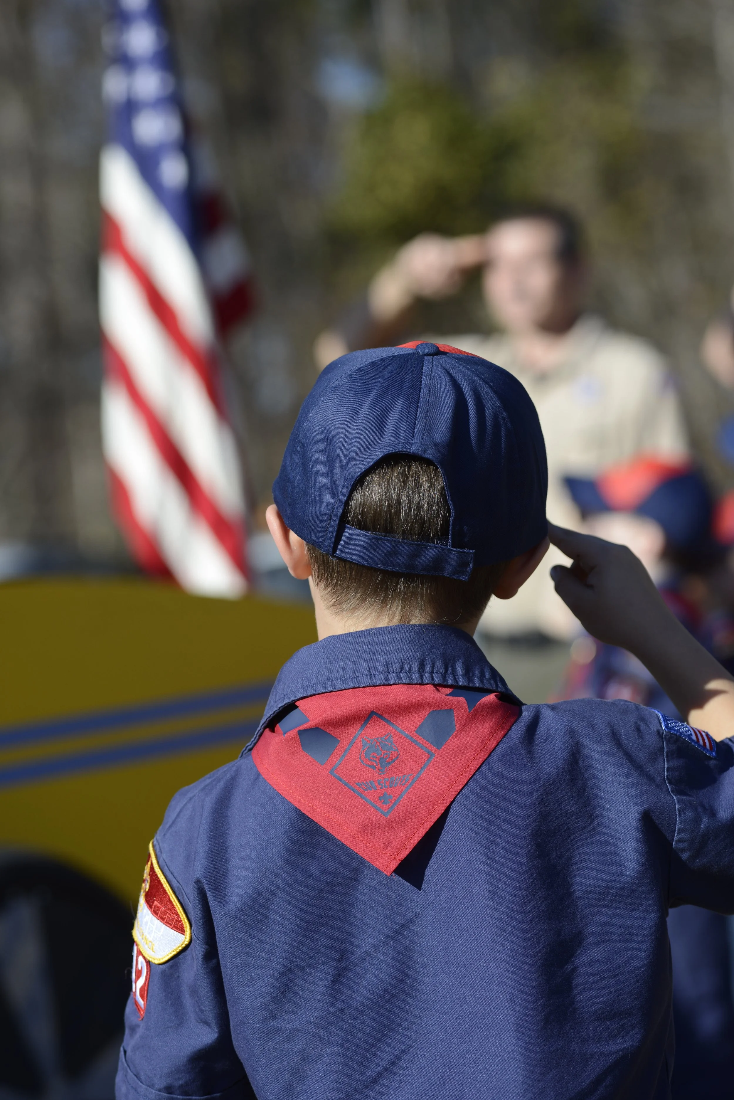 Boy in a navy Cub Scouts uniform and cap raising his hand at an outdoor event with an American flag in the background