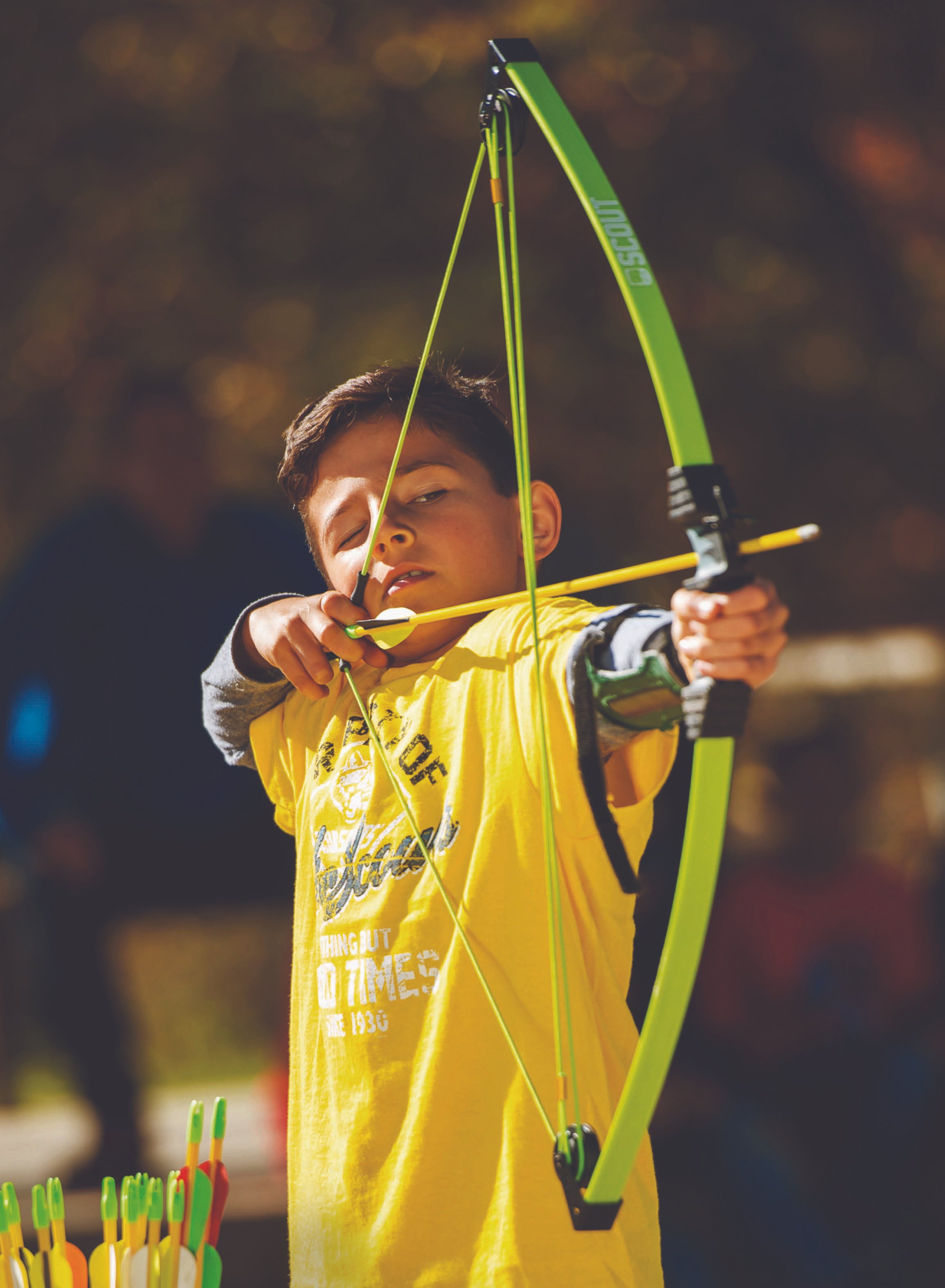 Young boy with brown hair, wearing a yellow shirt, aiming a small green and black toy bow and arrow outdoors. Blurred background with warm colors.