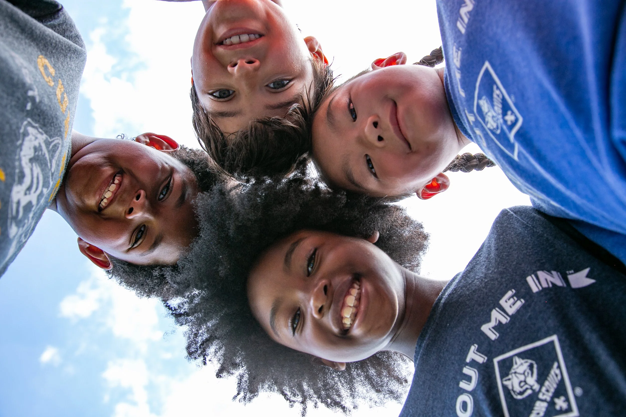 A group of children looking down at the camera, smiling with their faces close together, outdoors with a blue sky and clouds in the background.