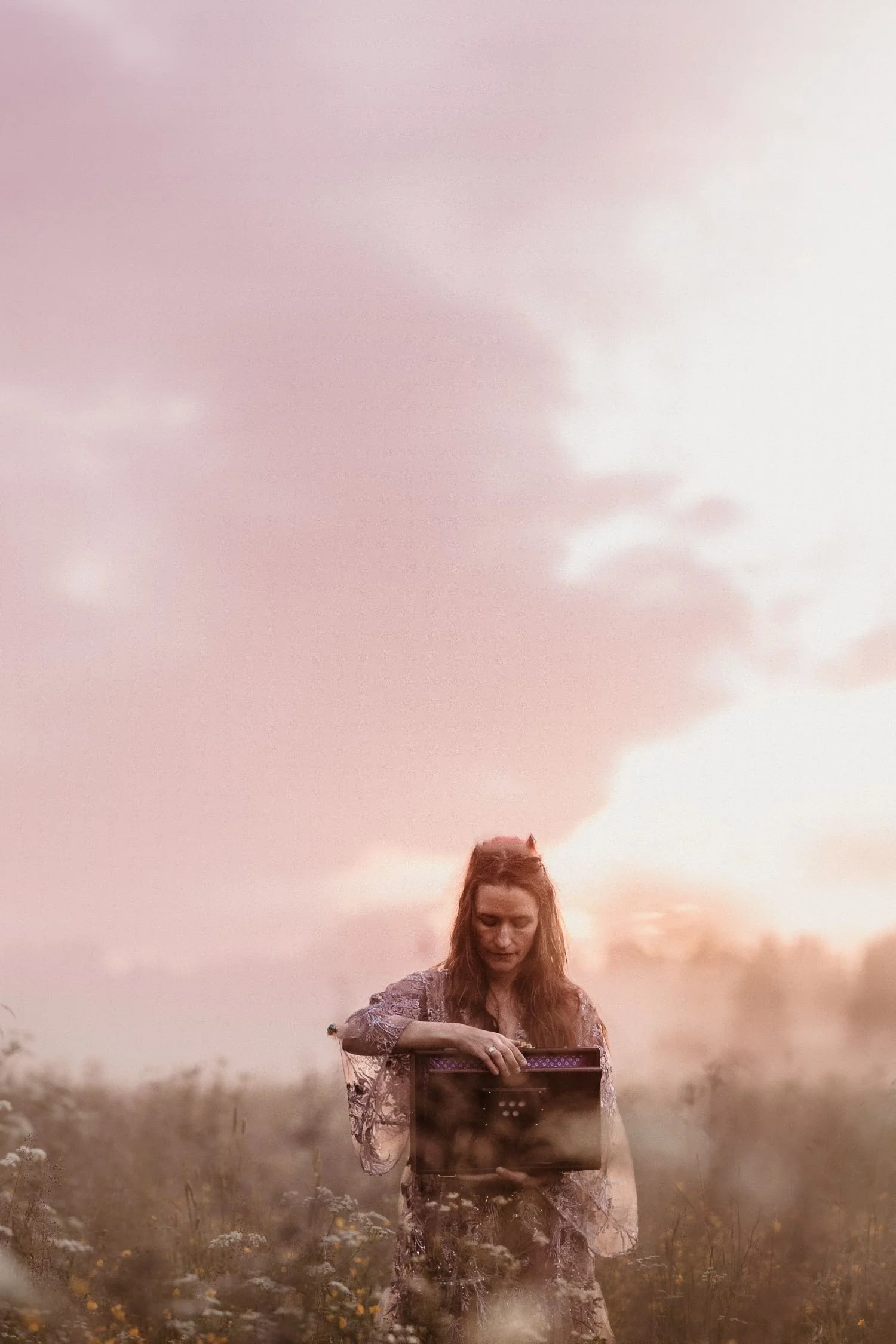 A woman with red hair in a dress standing outdoors in a field at sunset, holding and looking at a framed photograph or picture.