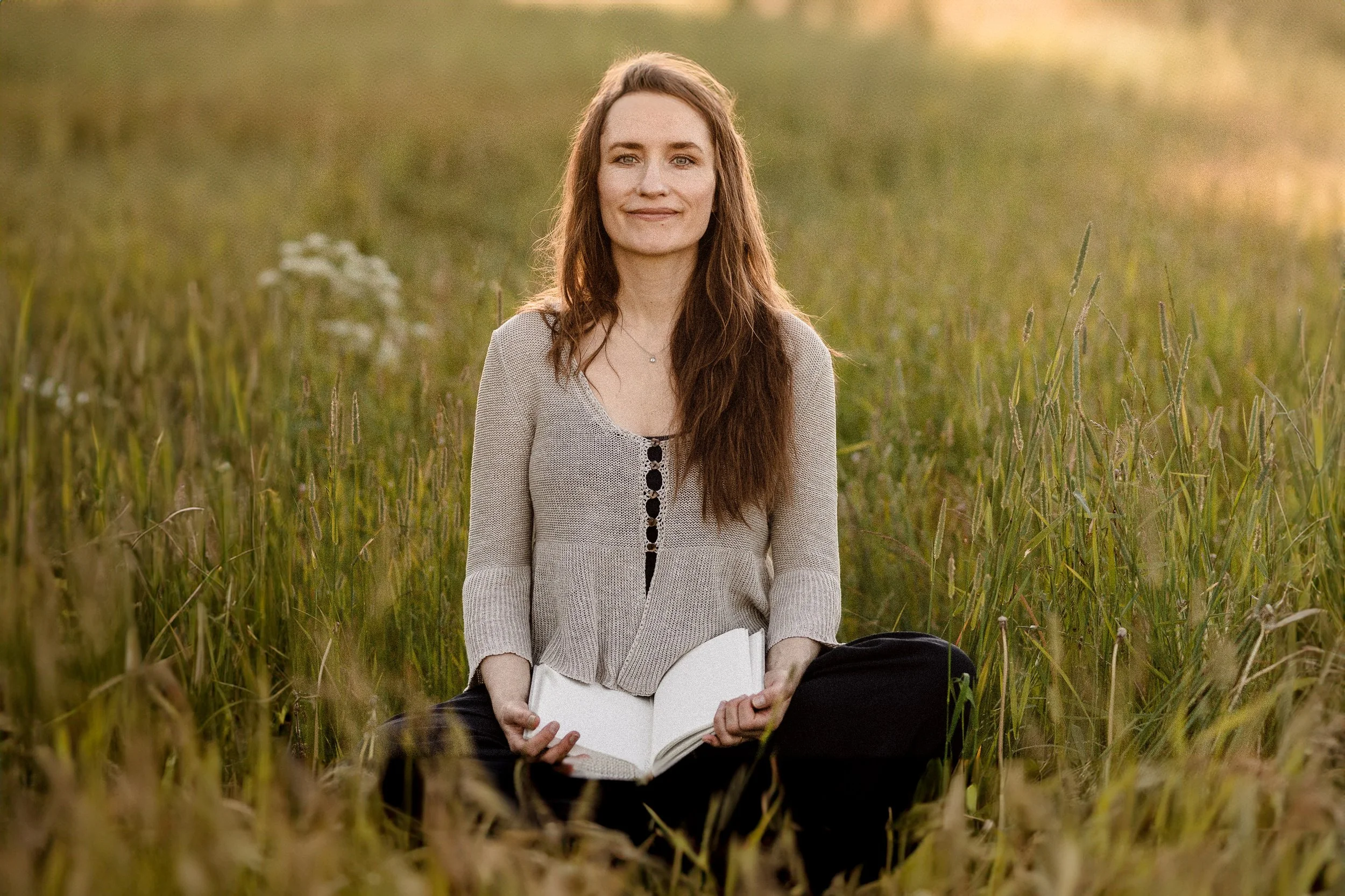A woman with long brown hair, wearing a beige cardigan, sitting in a field of tall grass at sunset, holding an open book.