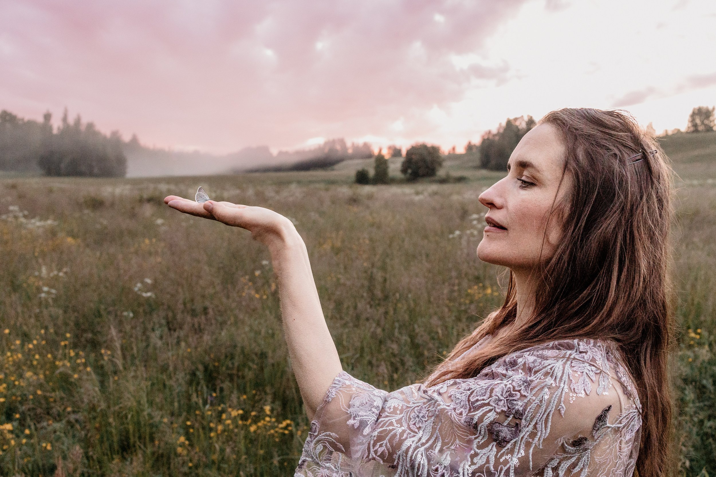 A woman with long brown hair holding a butterfly on her fingers outdoors at sunset in a grassy field.
