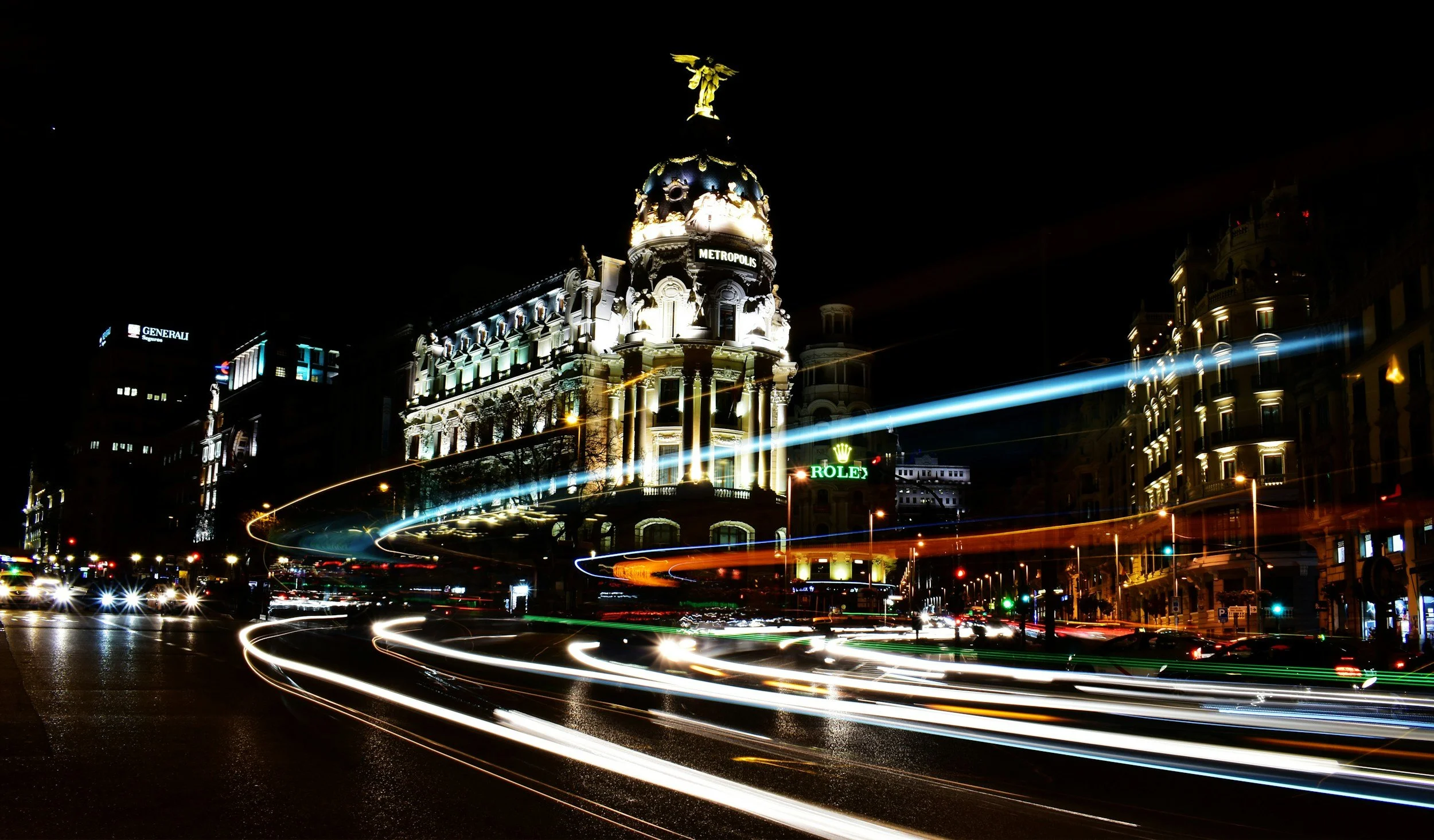 Night view of a historic building with a prominent dome topped with a statue, surrounded by illuminated modern buildings and busy street lights at night, with light trails from moving vehicles.