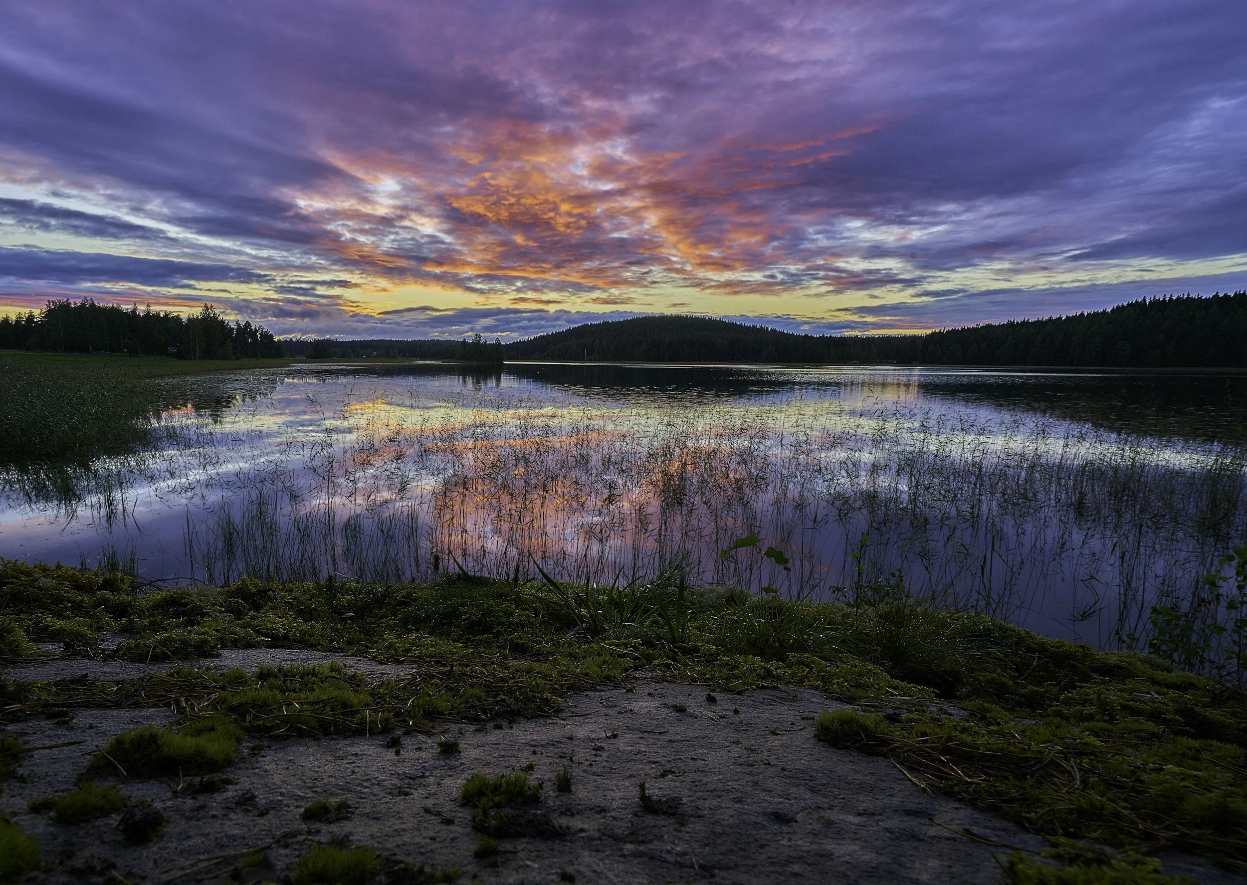 Sunset over a calm body of water with colorful clouds in the sky, surrounded by trees in the distance.
