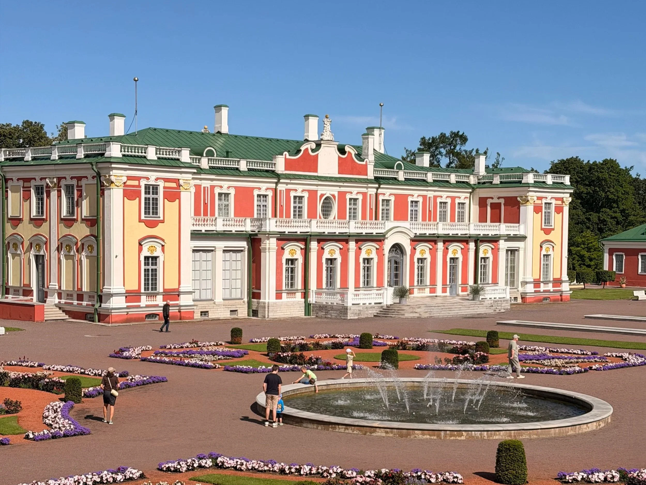 A large, ornate mansion with a green roof and pink and white exterior, surrounded by a landscaped garden with flower beds, a circular fountain, and people walking around on a sunny day with a clear blue sky.