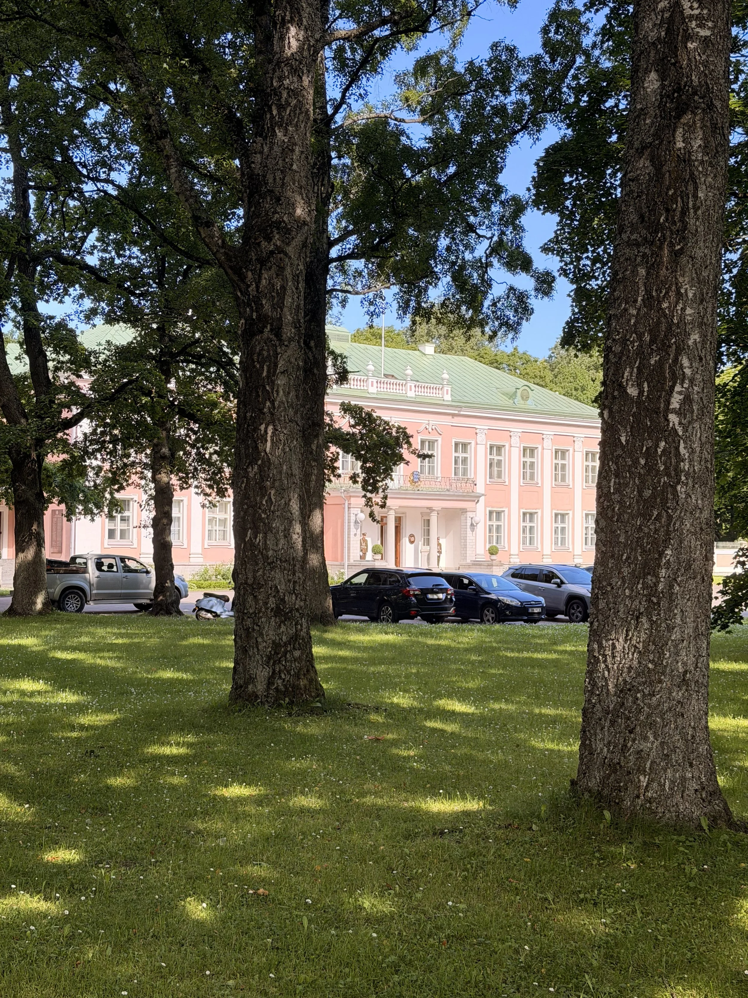 View of a pink historic building with white architectural accents, seen through large trees with green leaves in a park with green grass and a parking lot with cars.