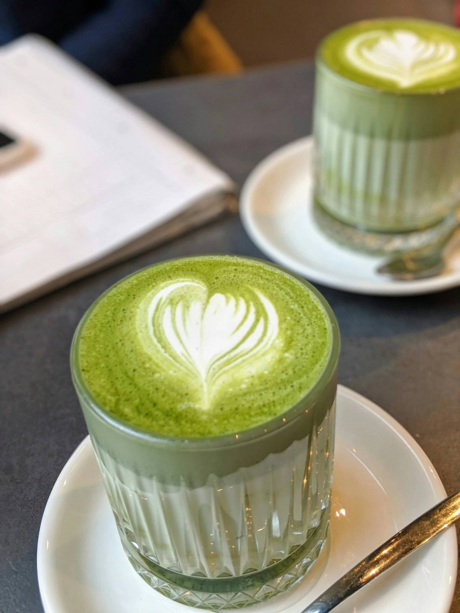 Un verre de matcha latte avec un motif de cœur en latte art, placé sur une assiette blanche, avec une cuillère argentée à côté.