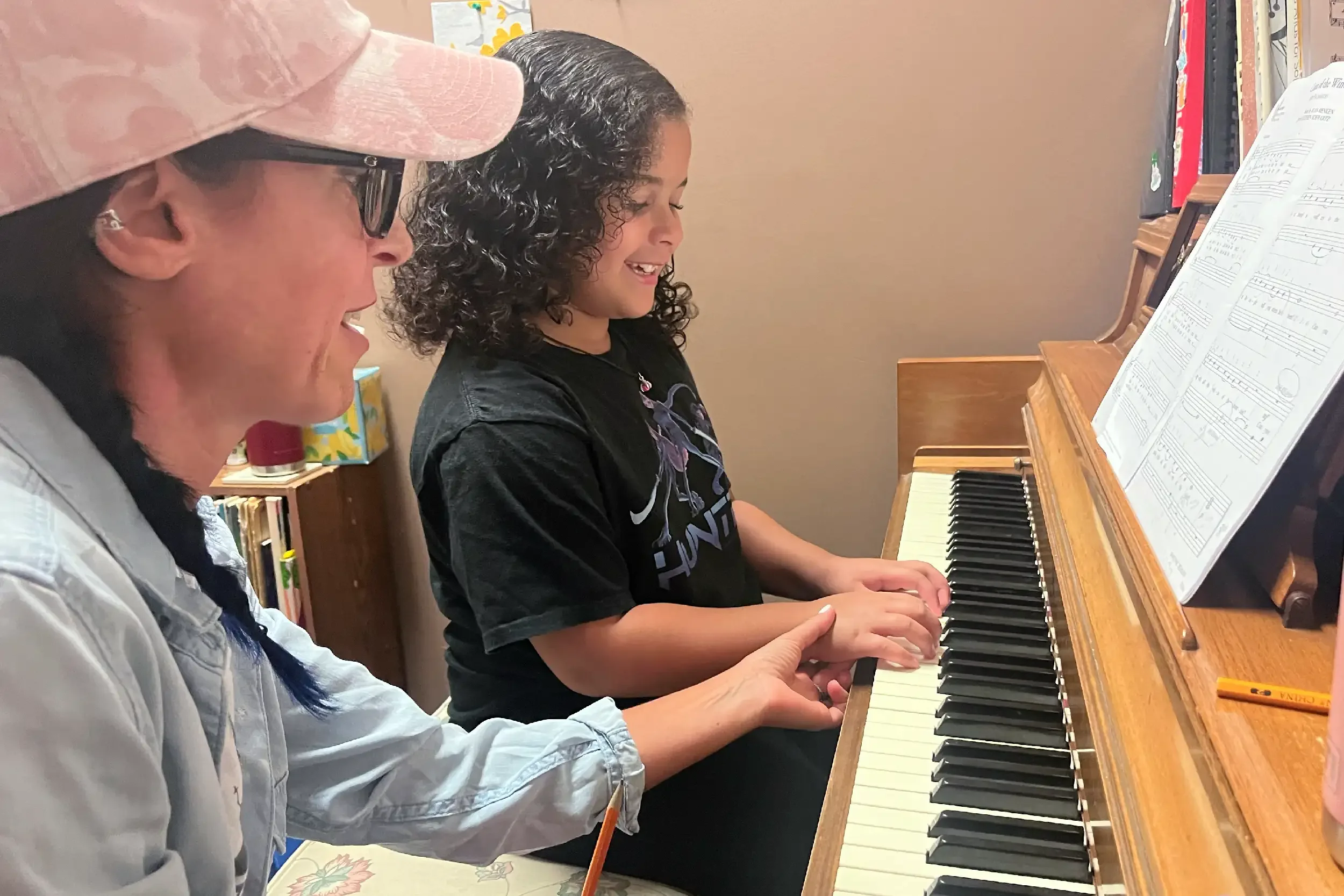 Two women sitting at a piano, smiling, with sheet music in front of them. One appears to be teaching or guiding the other while they play together.
