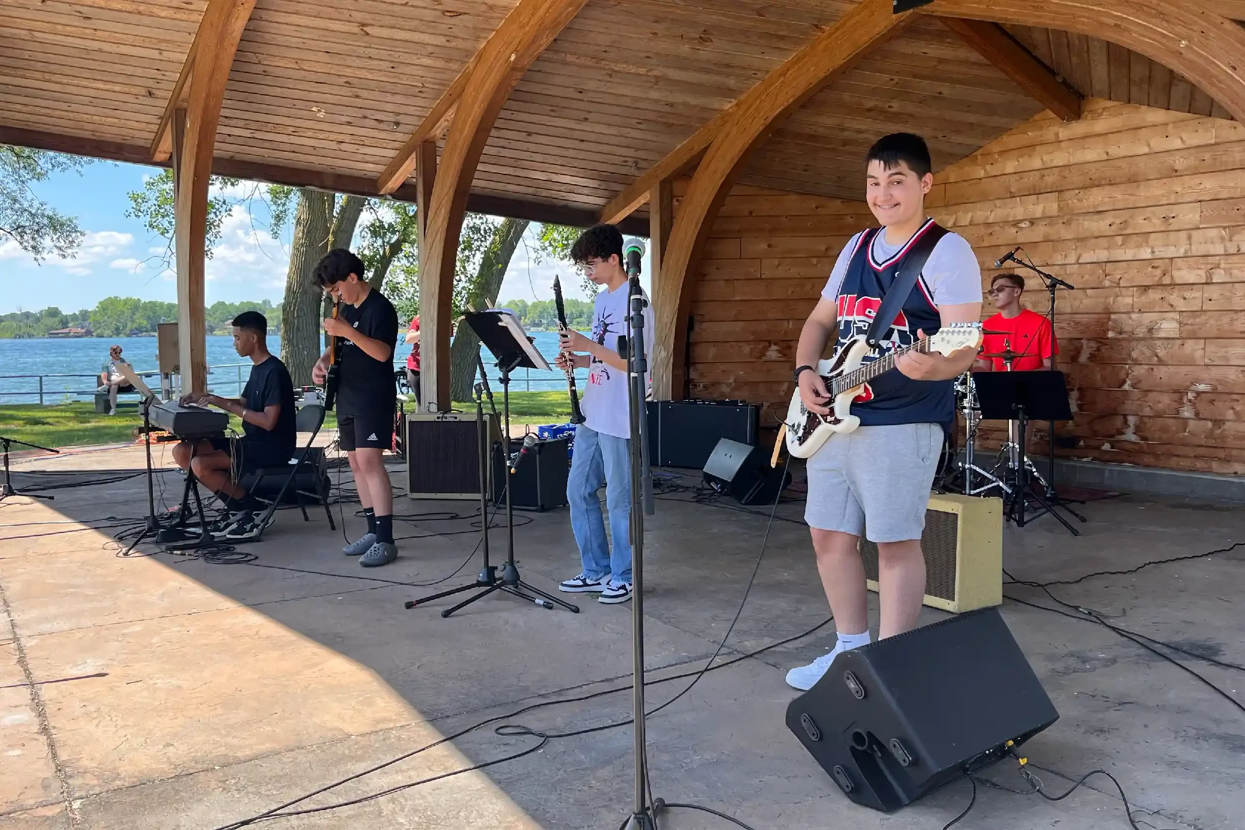 A group of young musicians performing on an outdoor stage near a lake, with one playing a guitar, two others playing keyboards and a saxophone, and a drummer in the background. There are spectators sitting and watching the performance.