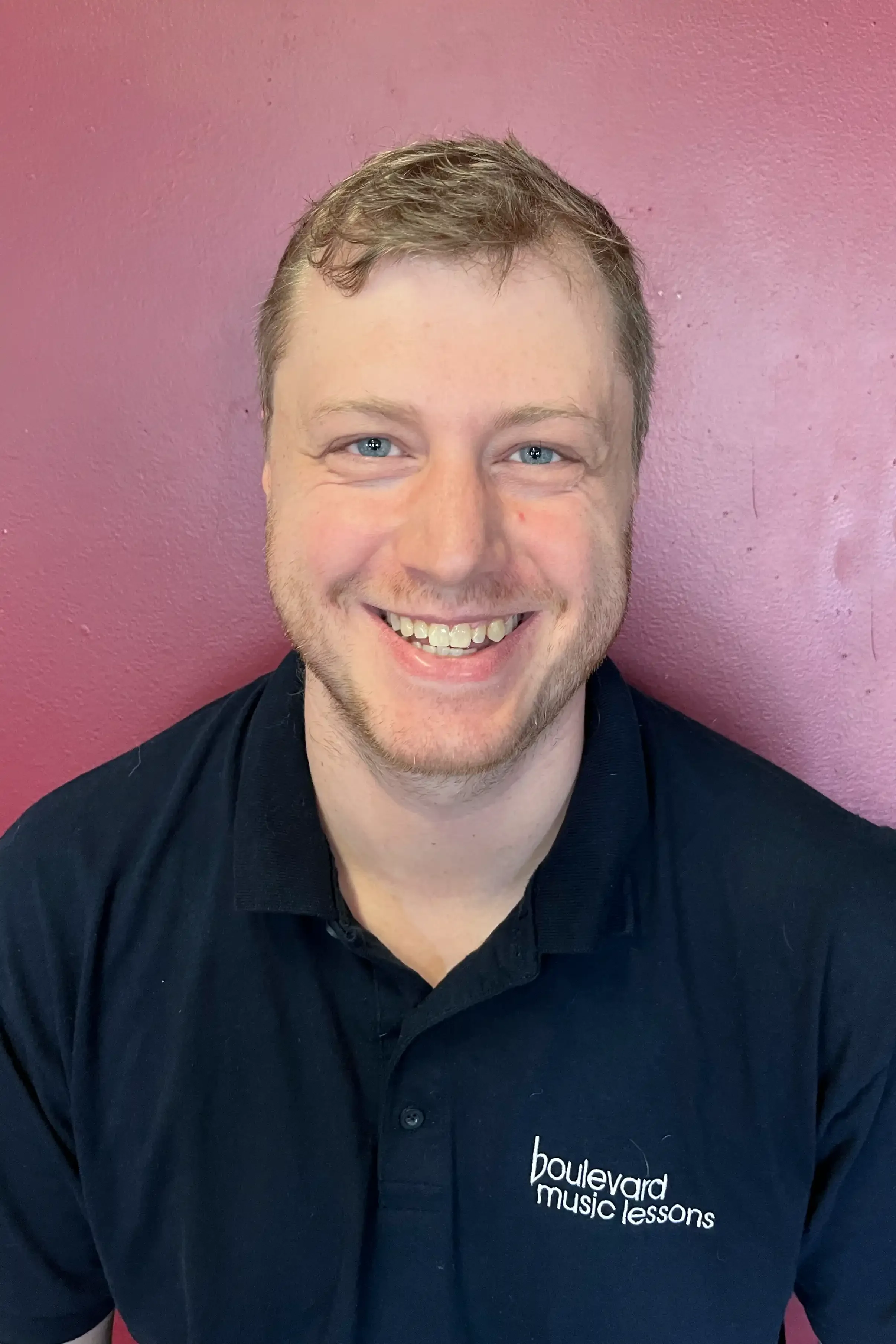 A smiling man with blue eyes, short light brown hair, wearing a black polo shirt with a logo that reads 'Boulevard Music Lessons,' standing against a pink wall.
