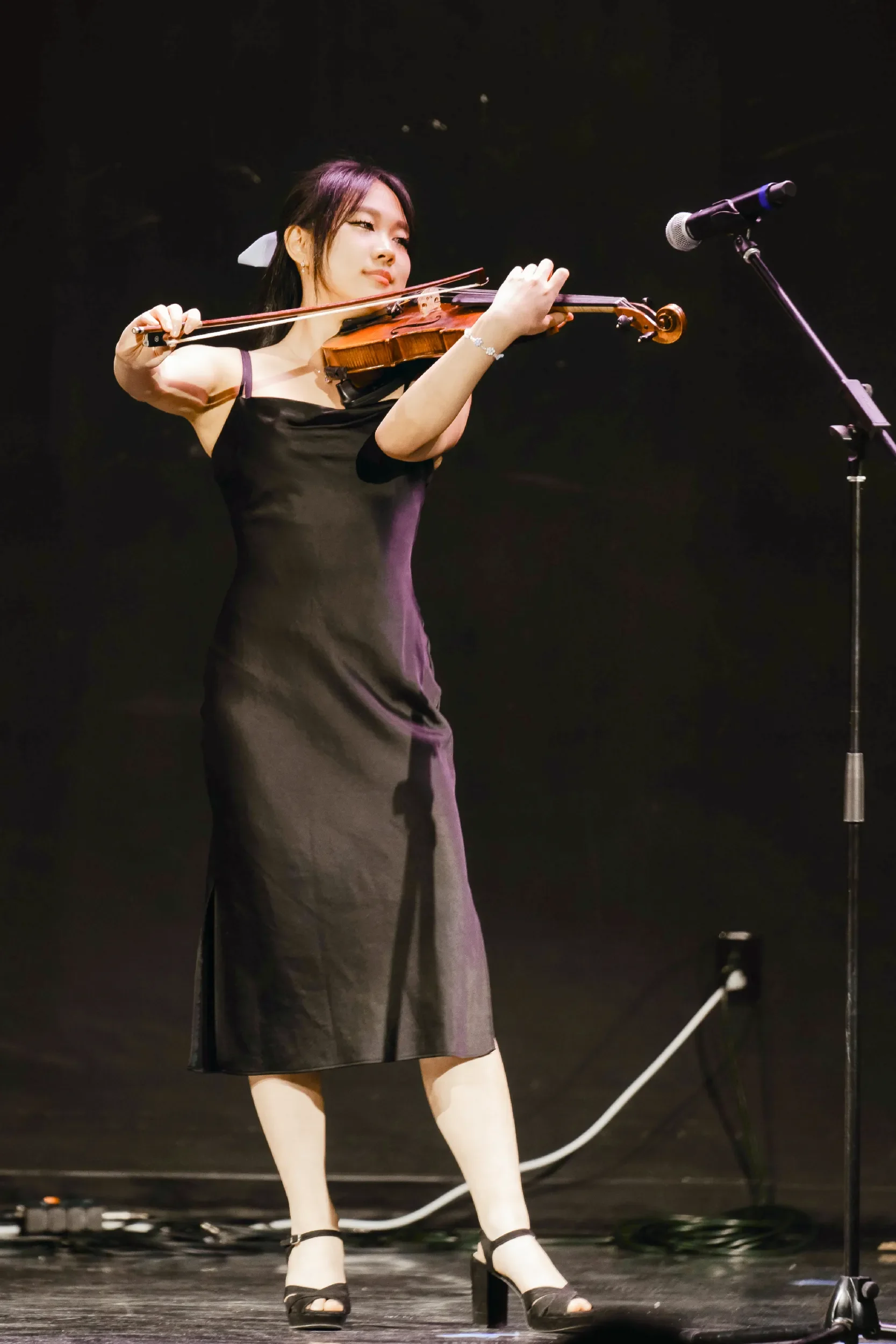 A woman in a black dress playing the violin on stage with a microphone nearby.