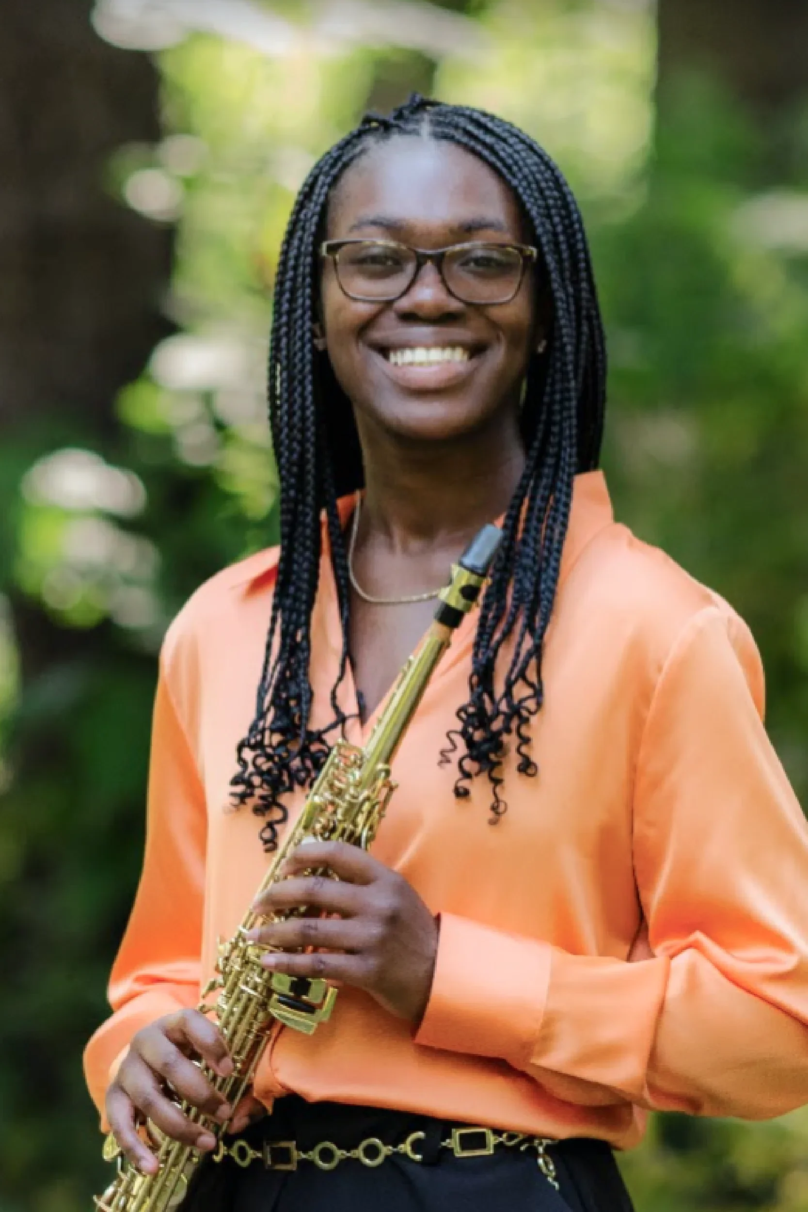 A smiling woman with braided hair and glasses, wearing an orange blouse and holding a flute, standing outdoors with a blurred green background.