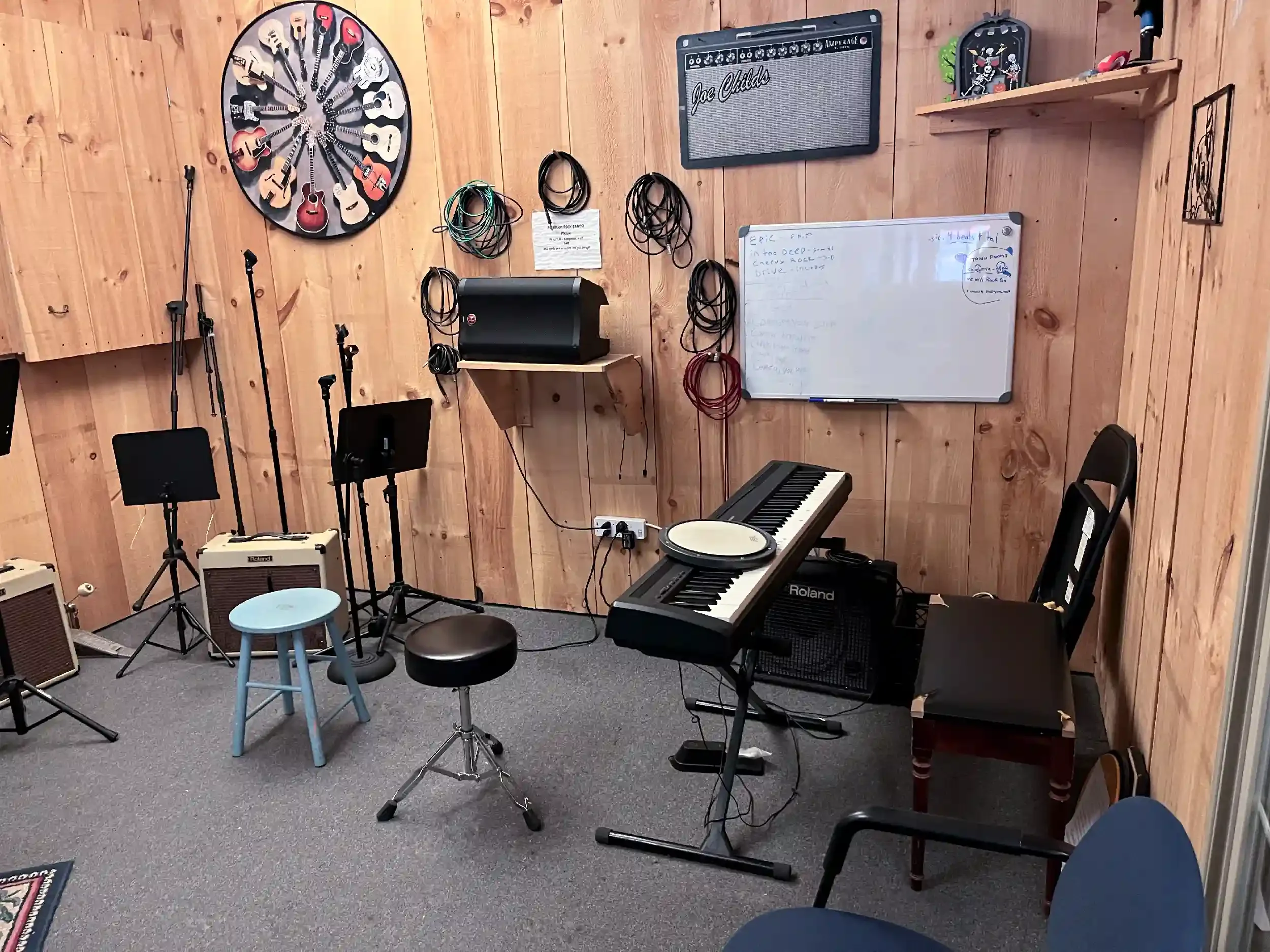 A music practice room with wooden walls, containing musical instruments, microphones, and recording equipment, including a keyboard, amplifier, and whiteboard.