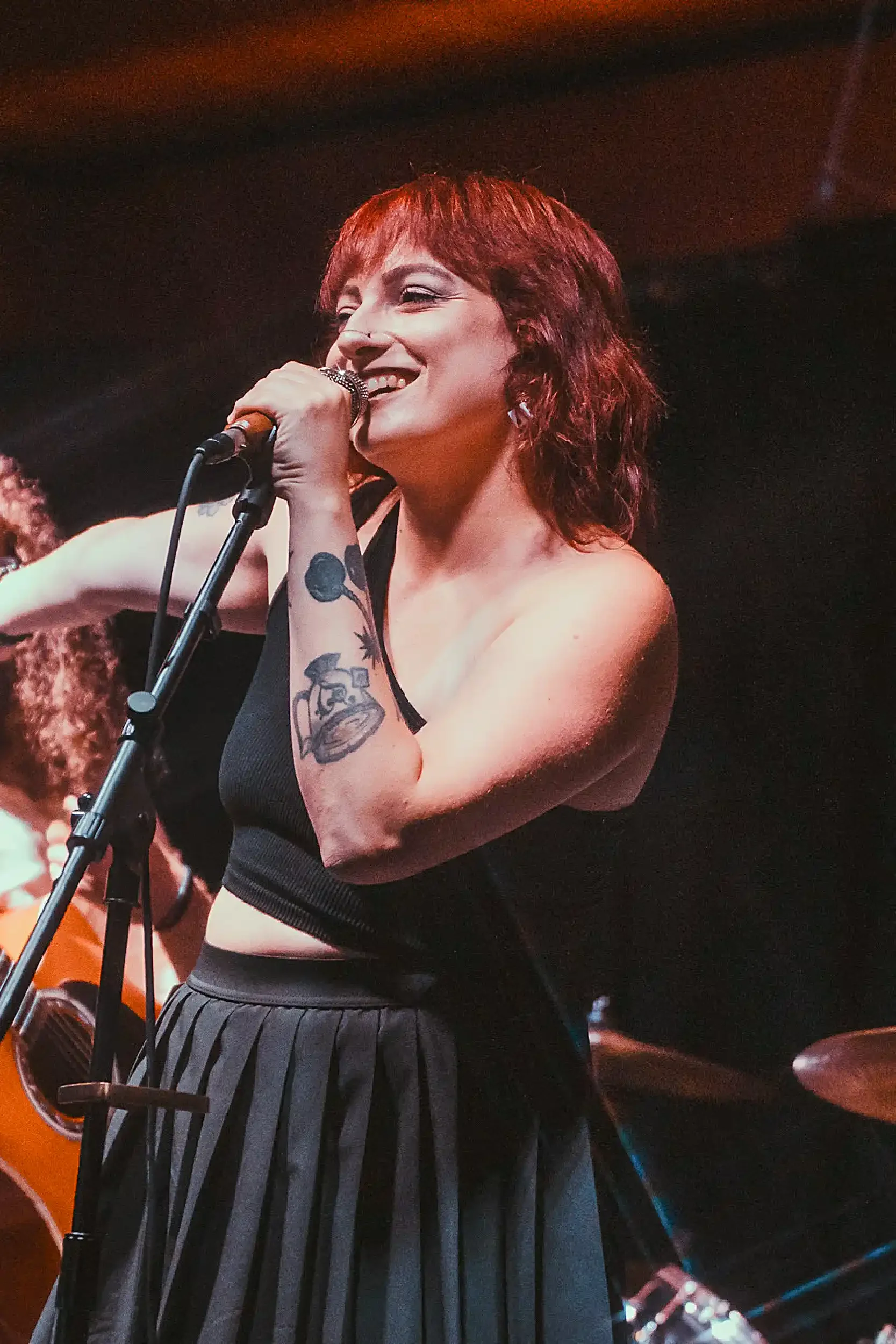 A woman with short red hair singing into a microphone on stage, wearing a black sleeveless top and pleated black skirt, with a tattoo on her left arm.