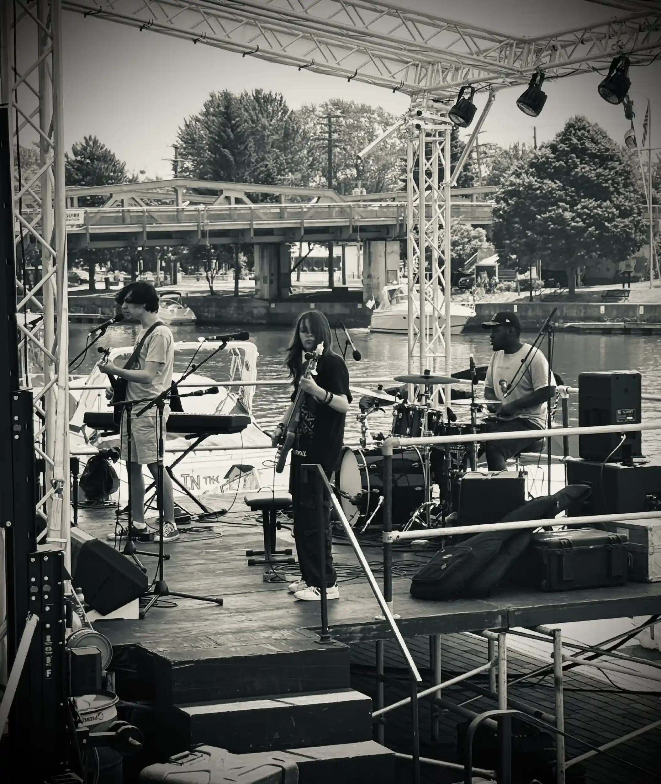 Musicians performing on an outdoor stage near a river, with trees and a bridge in the background, during daylight.