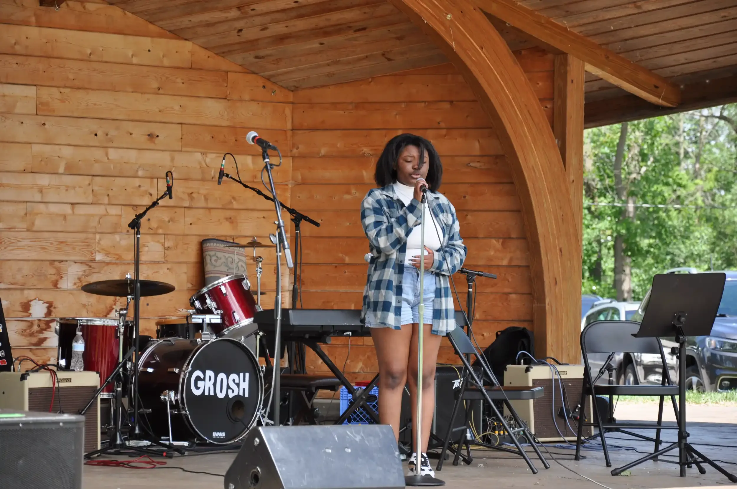 A young woman with dark hair, wearing a blue plaid shirt and shorts, singing into a microphone on a stage with musical instruments, including drums and a keyboard, in a wooden outdoor venue surrounded by trees.