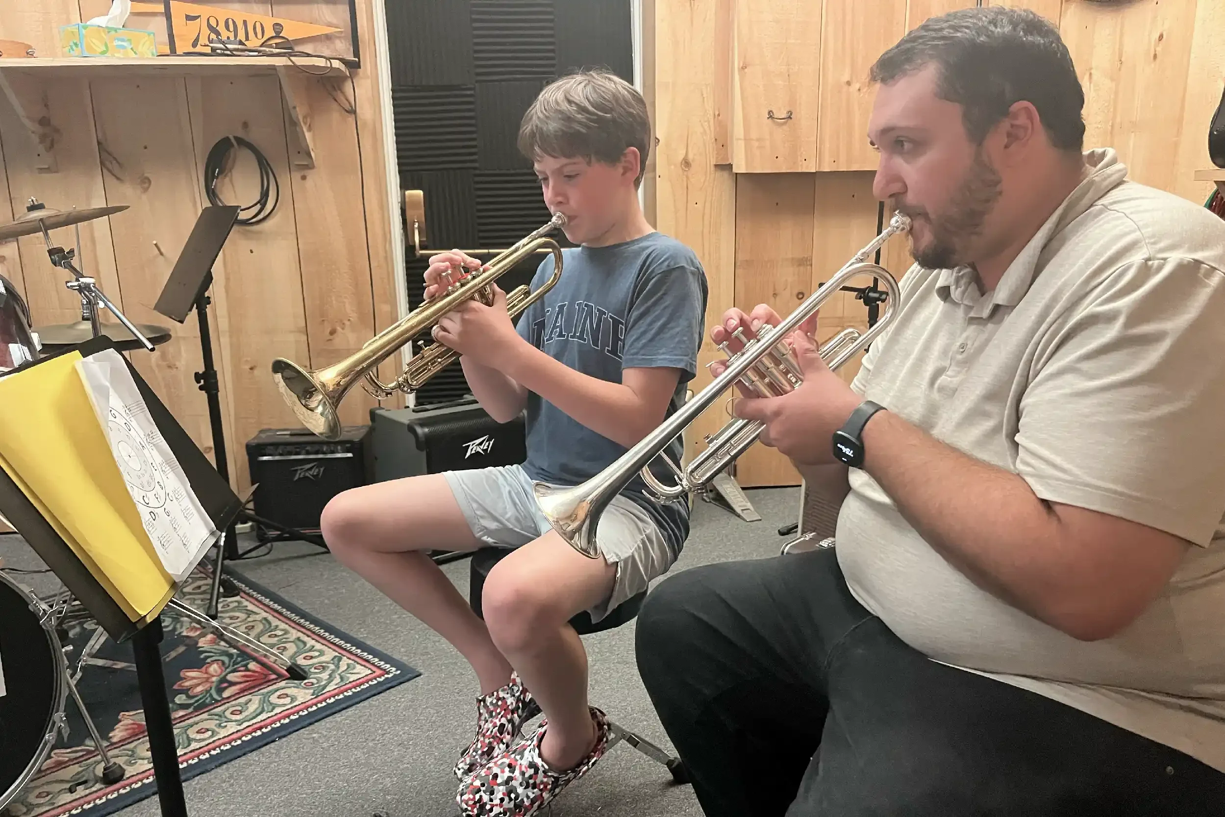 A young boy and an adult man playing trumpets in a music rehearsal room. The boy is sitting on a stool, reading sheet music, wearing a blue t-shirt, gray shorts, and patterned socks. The man is sitting next to him, wearing a beige polo shirt, and has