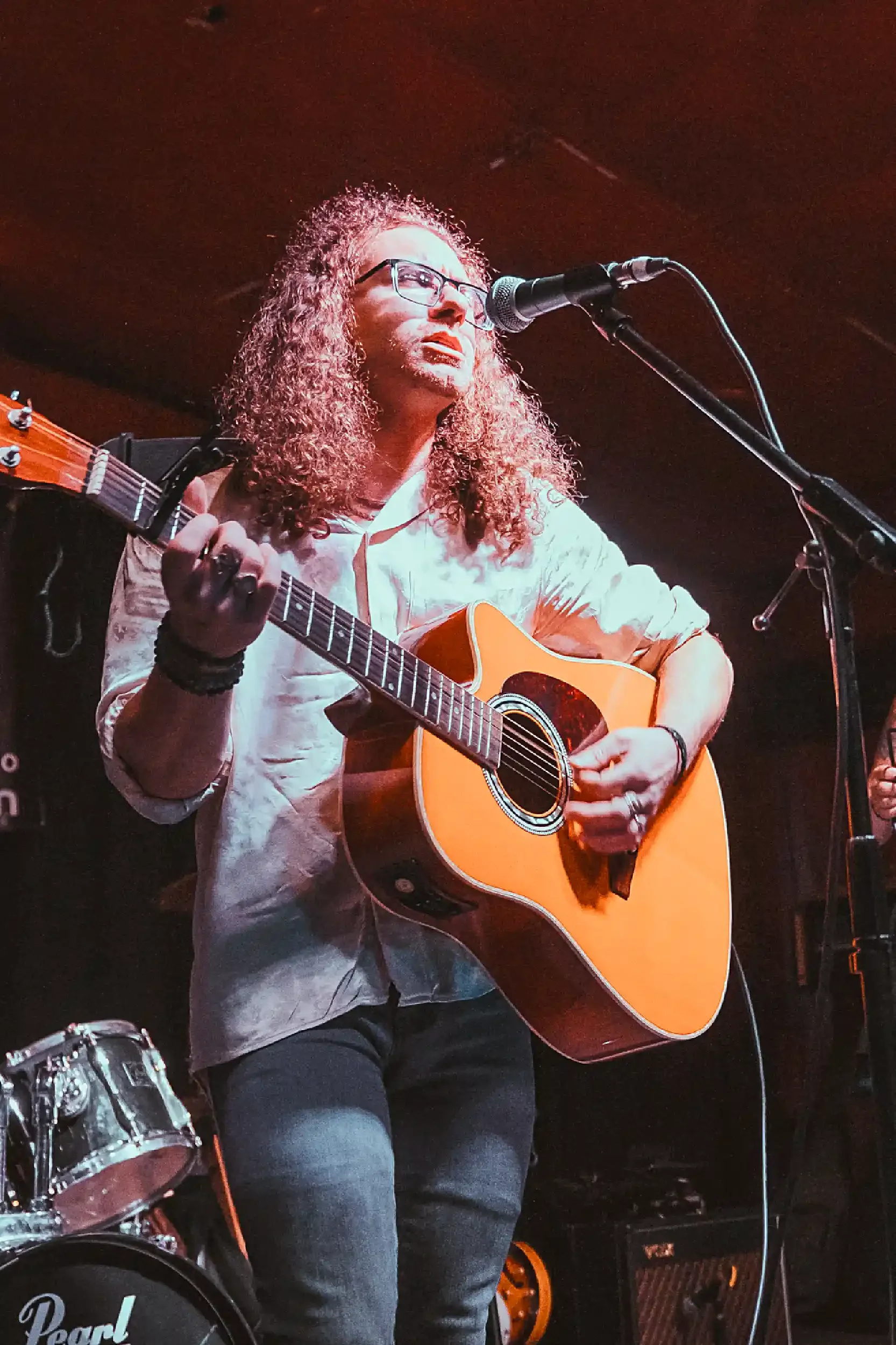 Musician with curly hair and glasses playing an acoustic guitar and singing into a microphone during a performance.