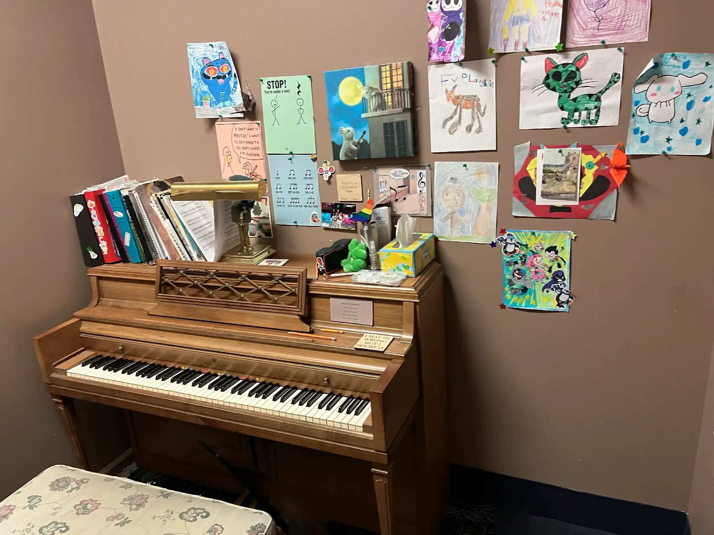 A wooden upright piano with a bookshelf on top, surrounded by children's drawings and artwork pinned to a beige wall. The piano keys are visible, and the room has a floral tablecloth in the foreground.
