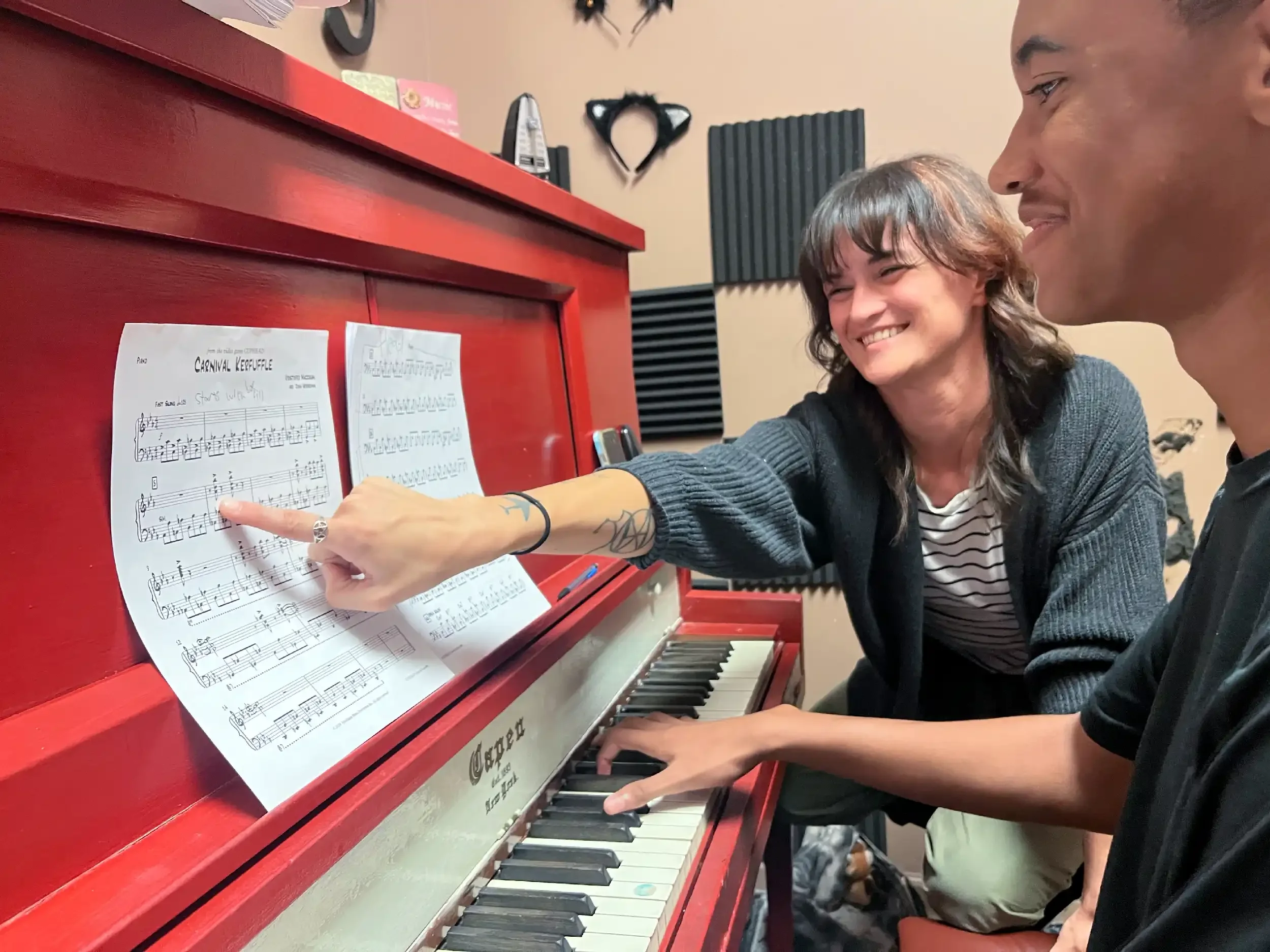 Two people, one with short brown hair and the other with longer gray hair, are sitting at a red piano. They are looking at and pointing to sheet music titled 'Carnival Rhapsody.' The person with gray hair is smiling, and the piano has black and white