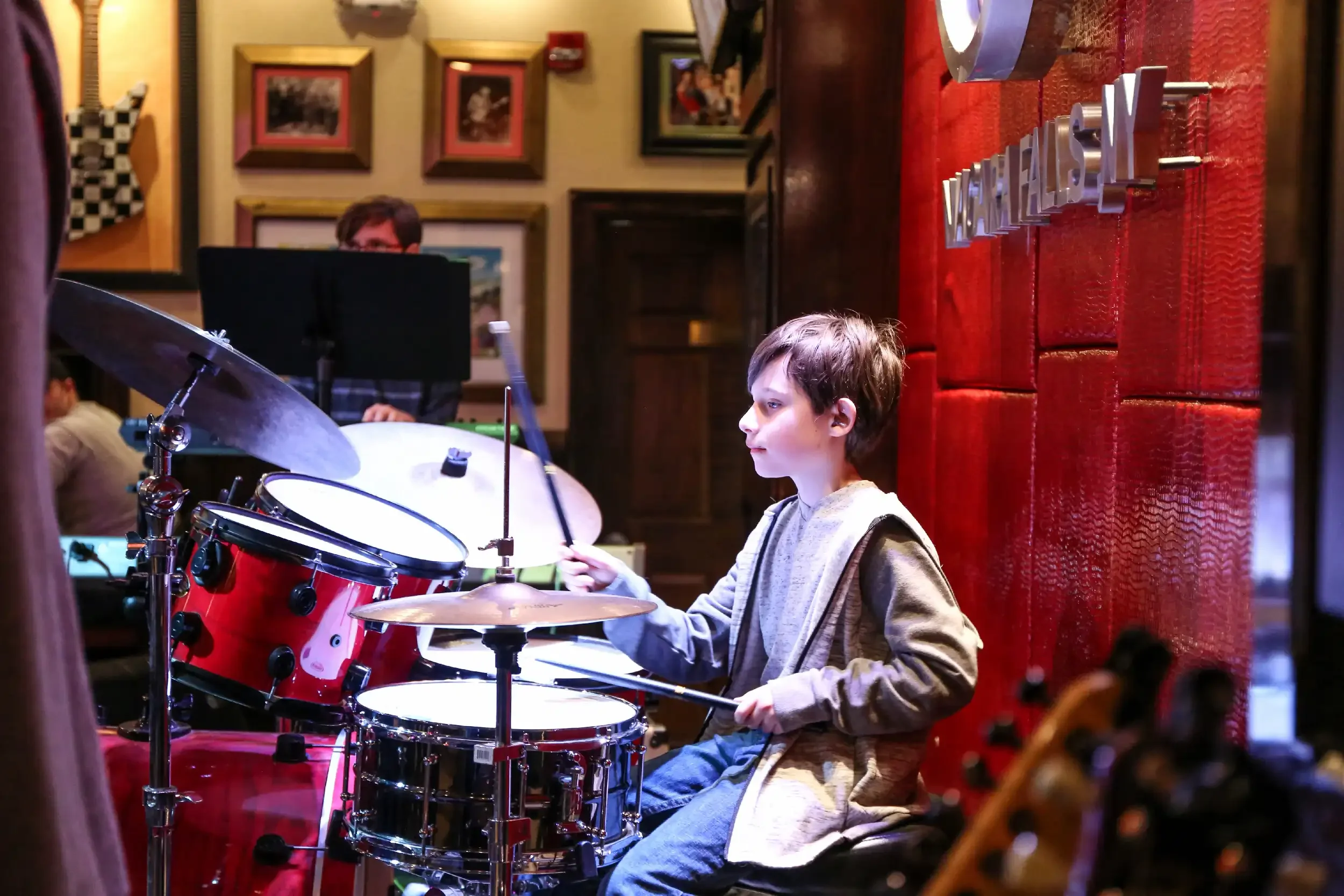 A young boy playing a red drum set in a cozy, warmly decorated room with framed pictures on the wall.