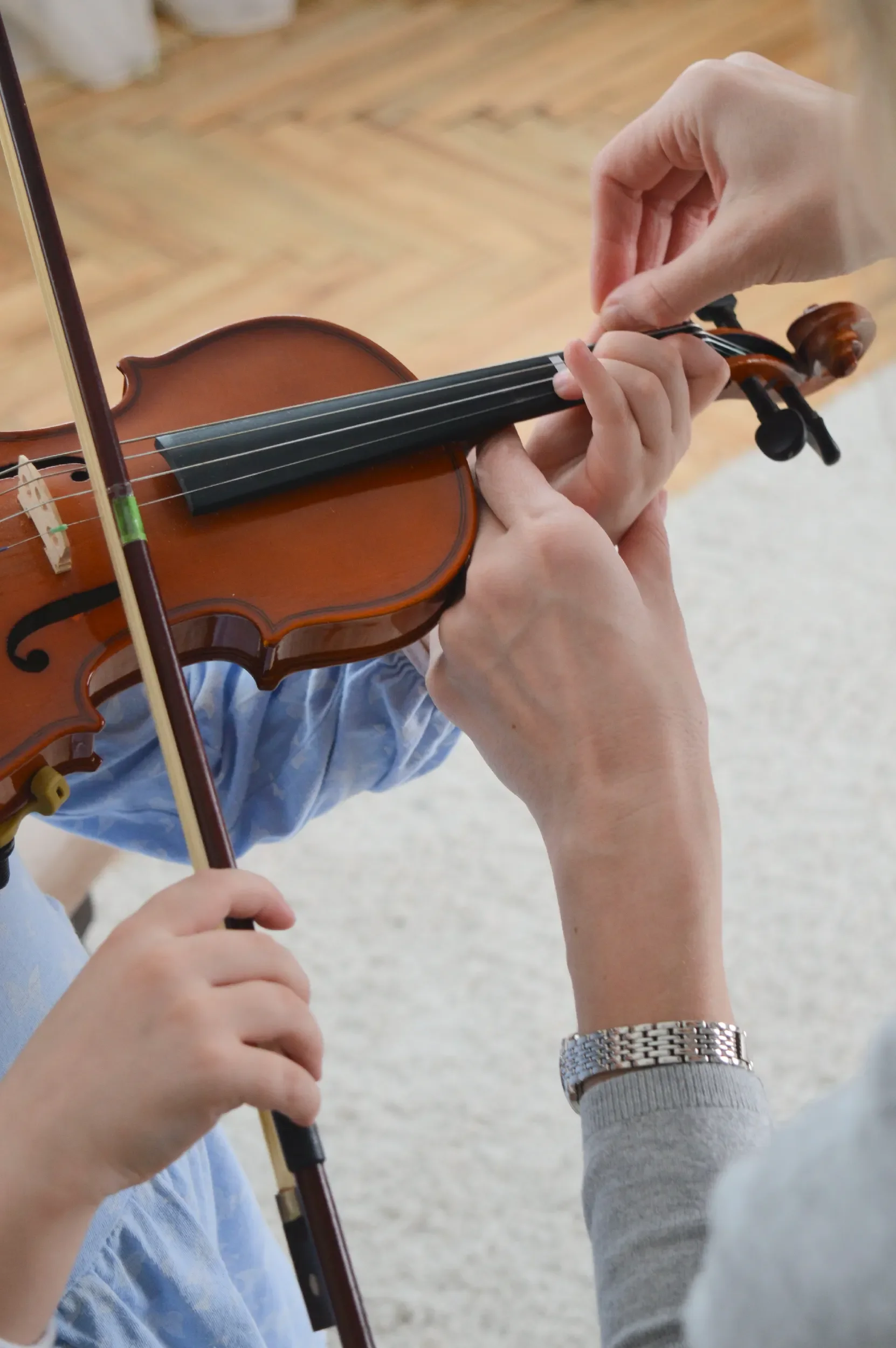 A person playing the violin while another person assists with the bow, in an indoor or outdoor setting with a wooden floor.