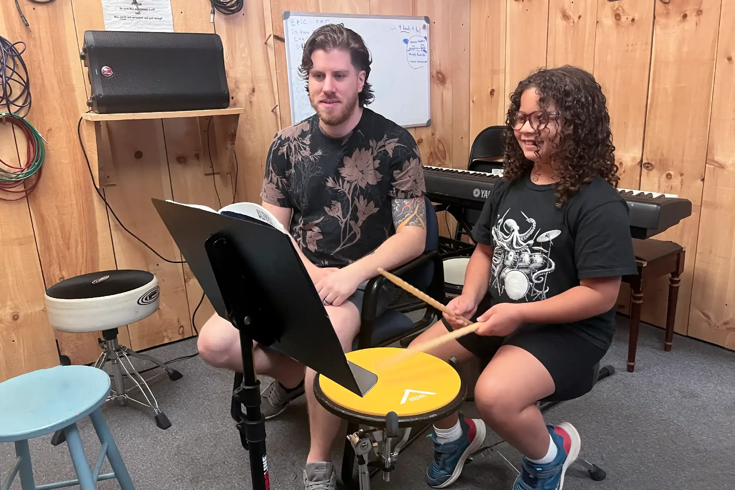 Two young men sitting in a music room, one playing drums and the other holding drumsticks. The room has wooden walls, a keyboard, a whiteboard, and musical equipment.