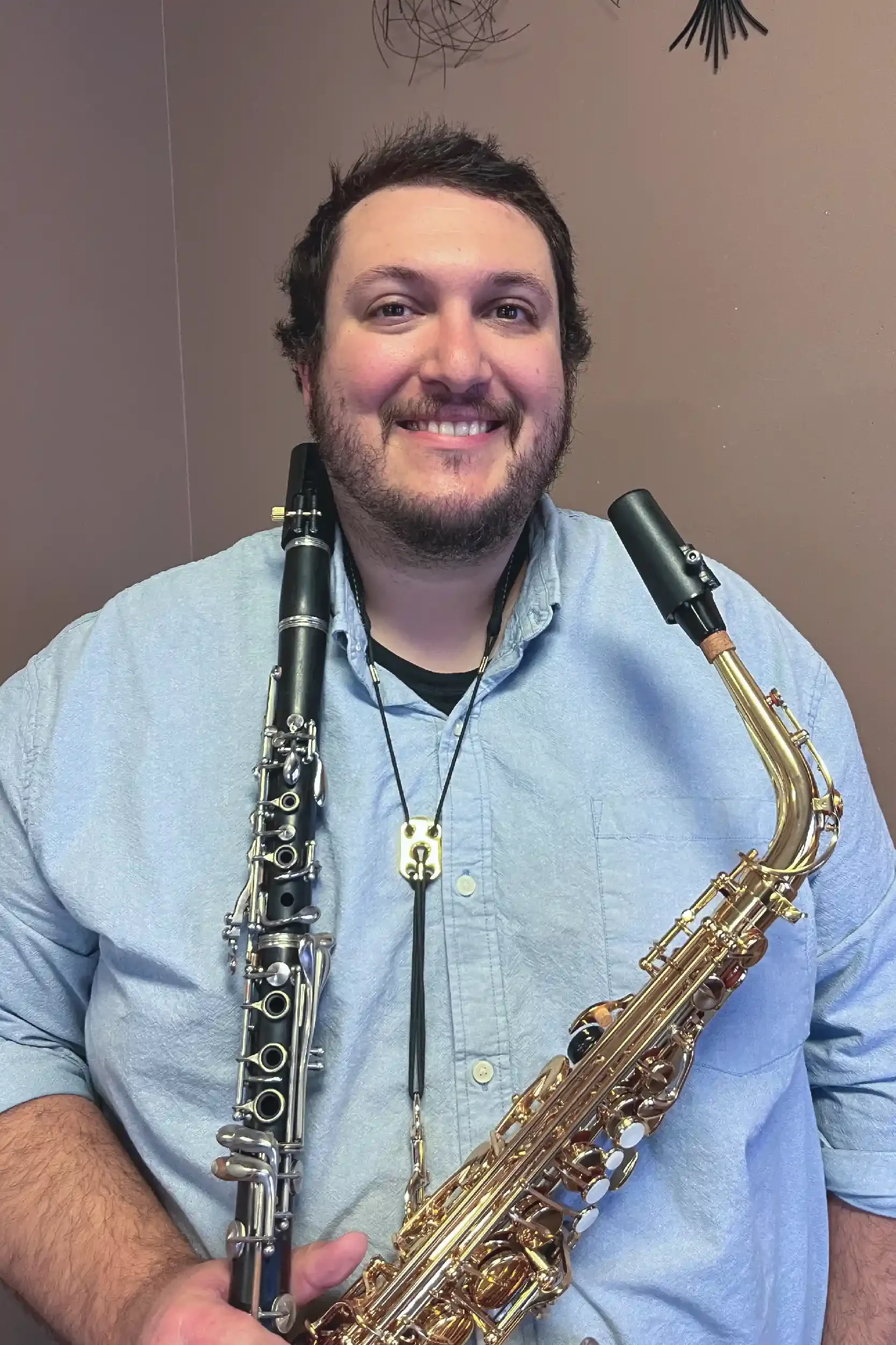 A smiling man with a beard holding a clarinet in his left hand and a saxophone in his right hand, both instruments resting on his shoulders, against a plain background.