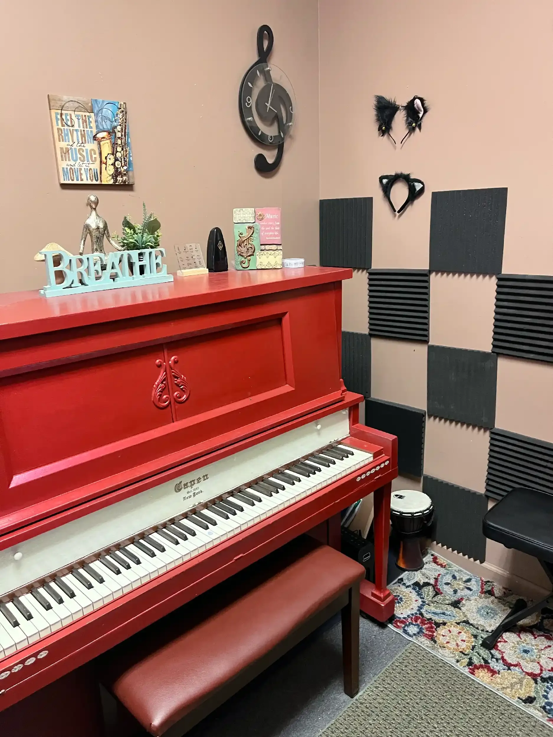 A red upright piano with a matching stool, a decorative metal figure, and a small potted plant on top. The piano is in a room with soundproof foam panels on the walls, a patterned rug on the floor, and a drum in the corner.