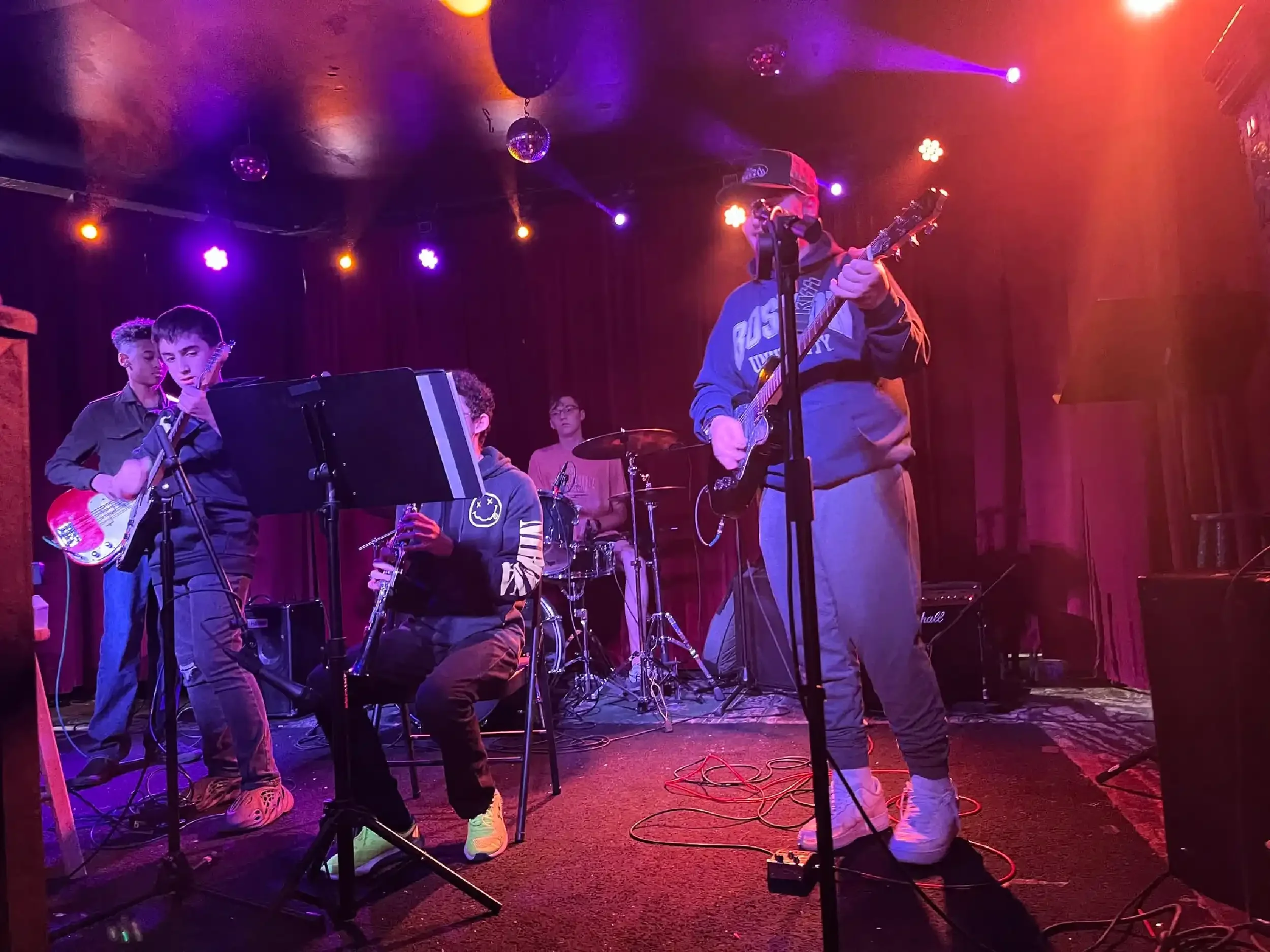 A music band performing on stage with a guitarist, a keyboard player, a drummer, and a singer. The stage is lit with purple and orange lights.