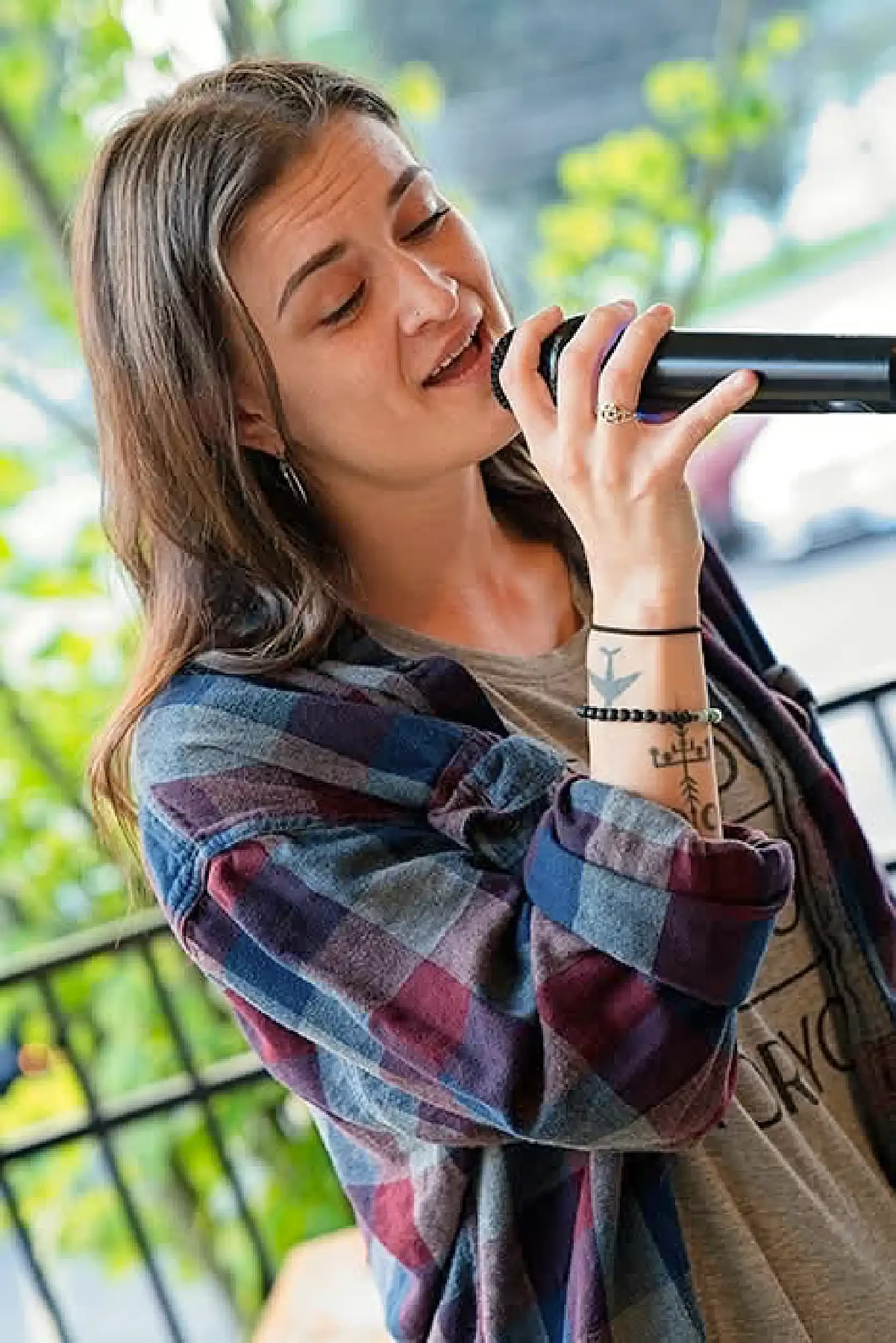 A young woman with long brown hair, wearing a plaid shirt and tattoos on her arm, is smiling and holding a black water bottle near her mouth, standing outdoors with a blurred background of greenery and a metal railing.