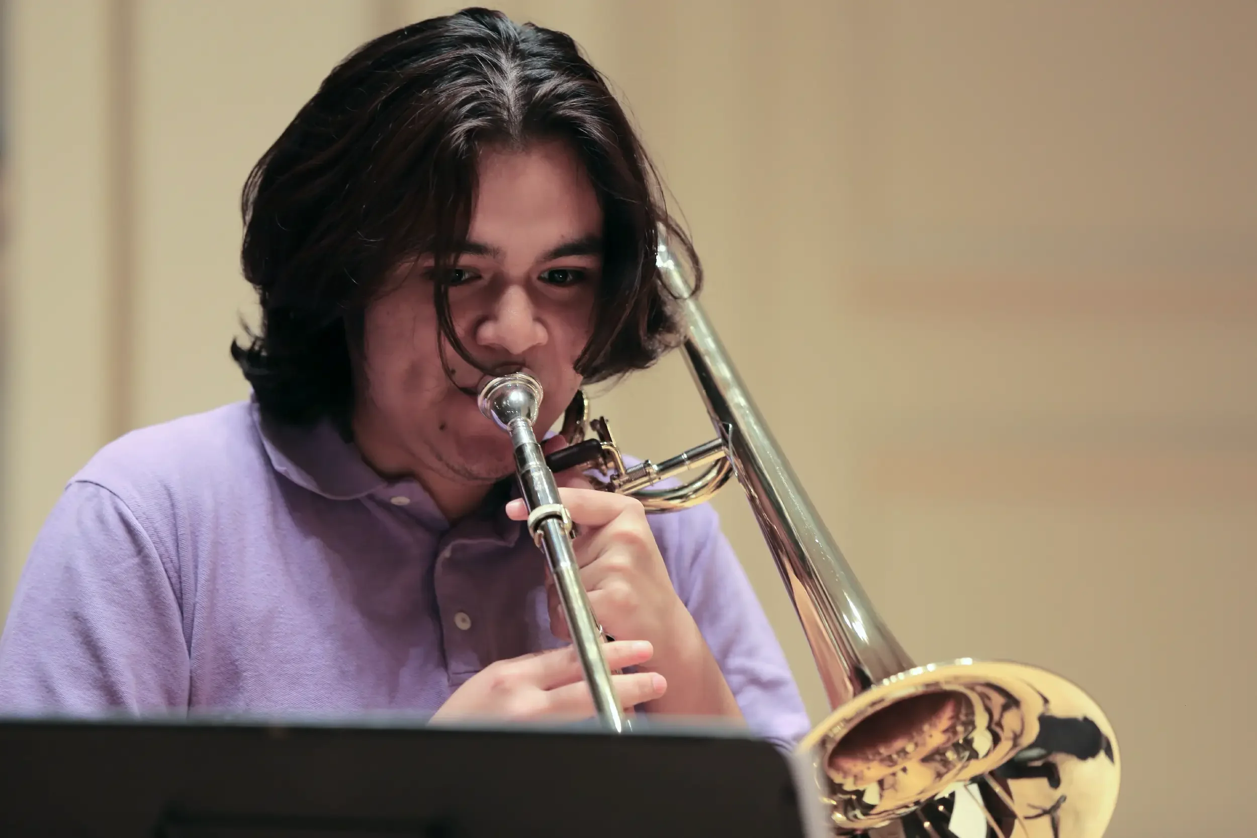 Young man with medium-length dark hair playing the trombone, wearing a lavender shirt, in a music studio.