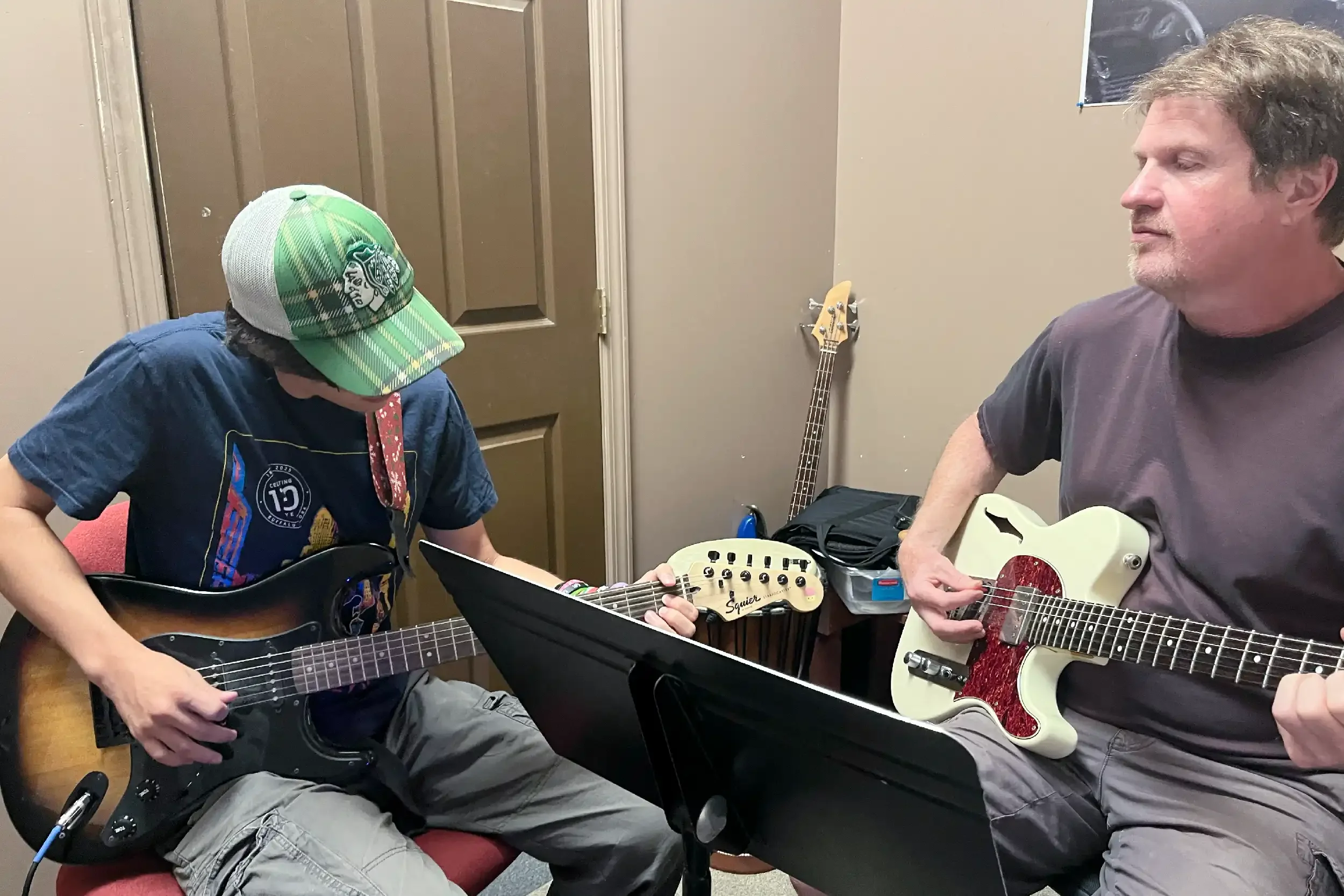 Two people playing acoustic guitars in a room, with a bass guitar propped against the wall behind them. One person is wearing a green and gray cap, while the other has curly gray hair.