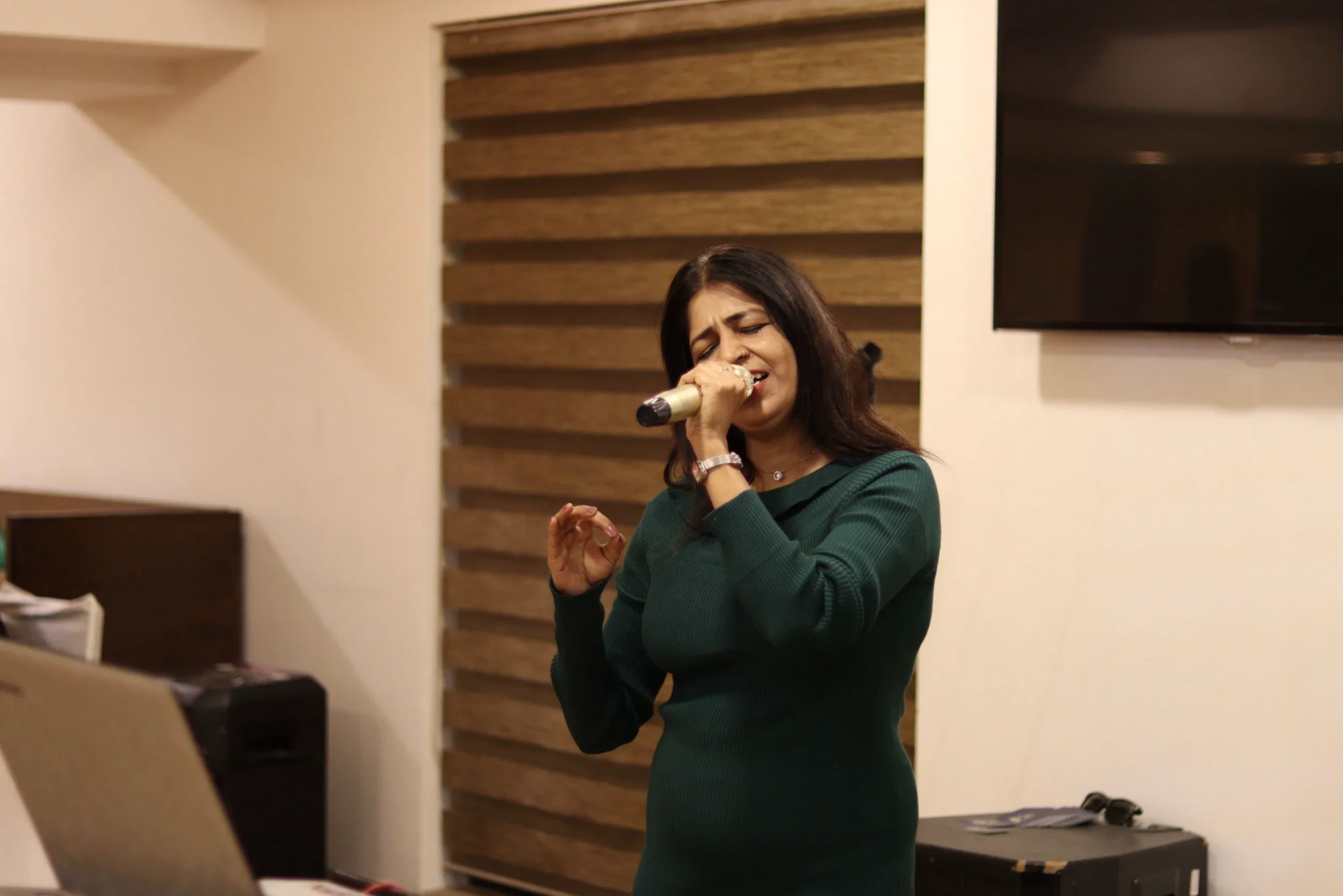 A woman singing passionately into a microphone in a room with wooden wall decor and a black flat-screen TV on the wall.