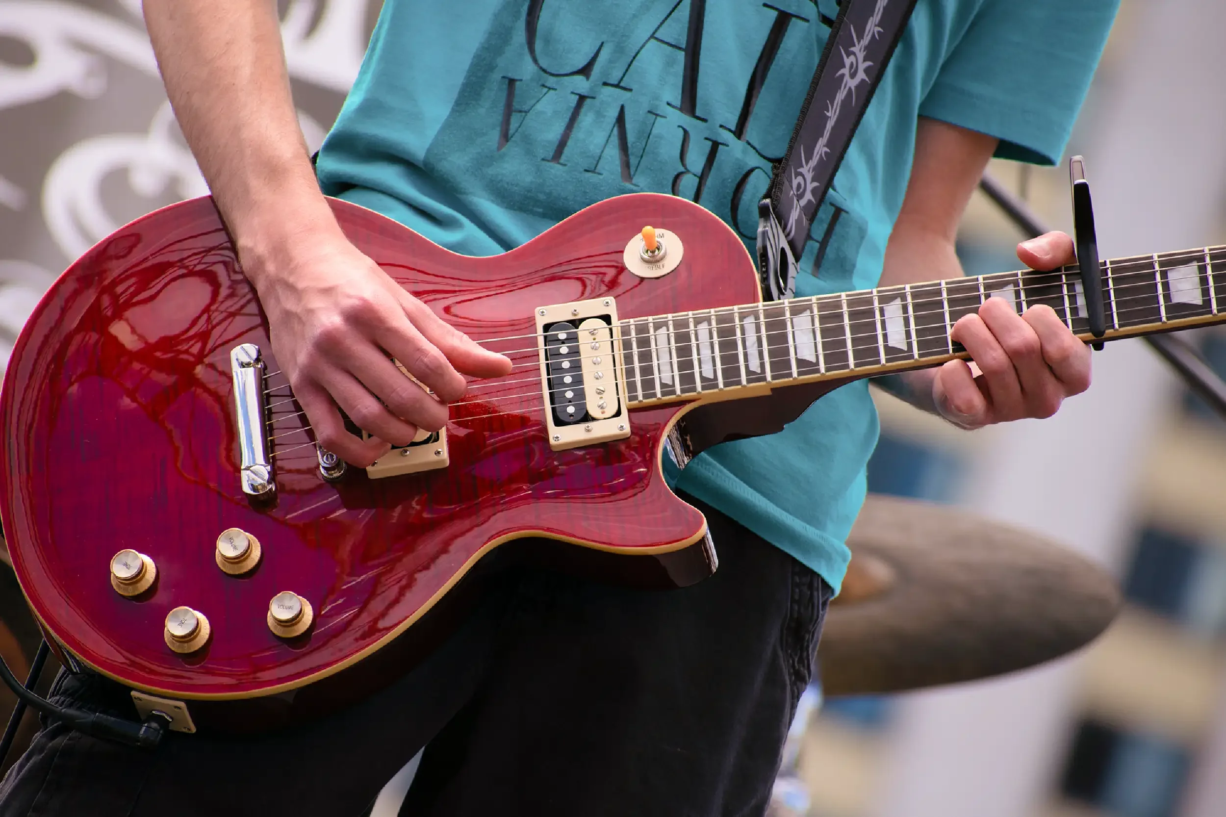 A person playing a red electric guitar outdoors, wearing a light blue T-shirt with black text and a black strap with a purple floral pattern.