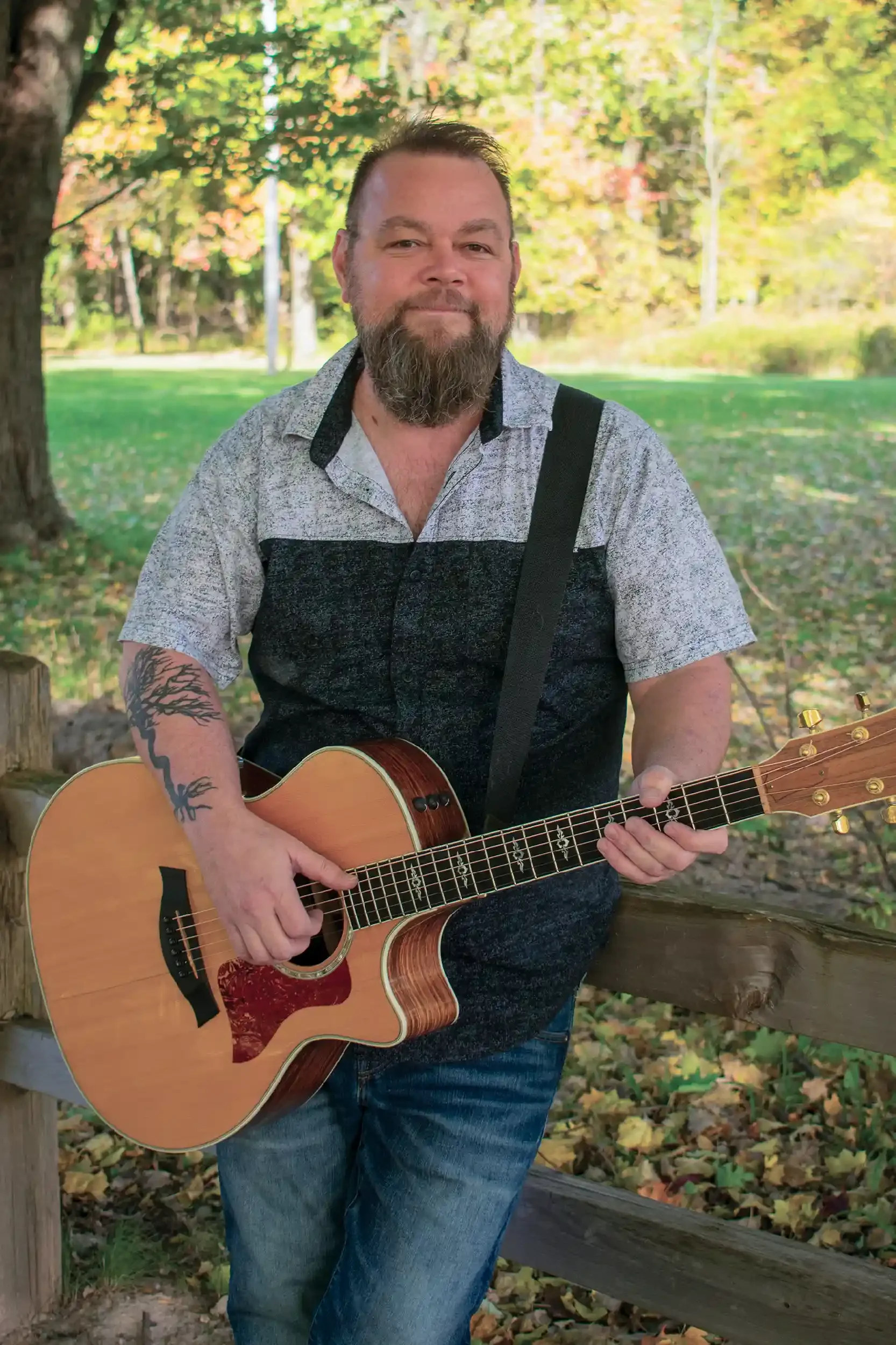 A man with a beard and tattoos playing an acoustic guitar outdoors in a park with autumn leaves and trees in the background.