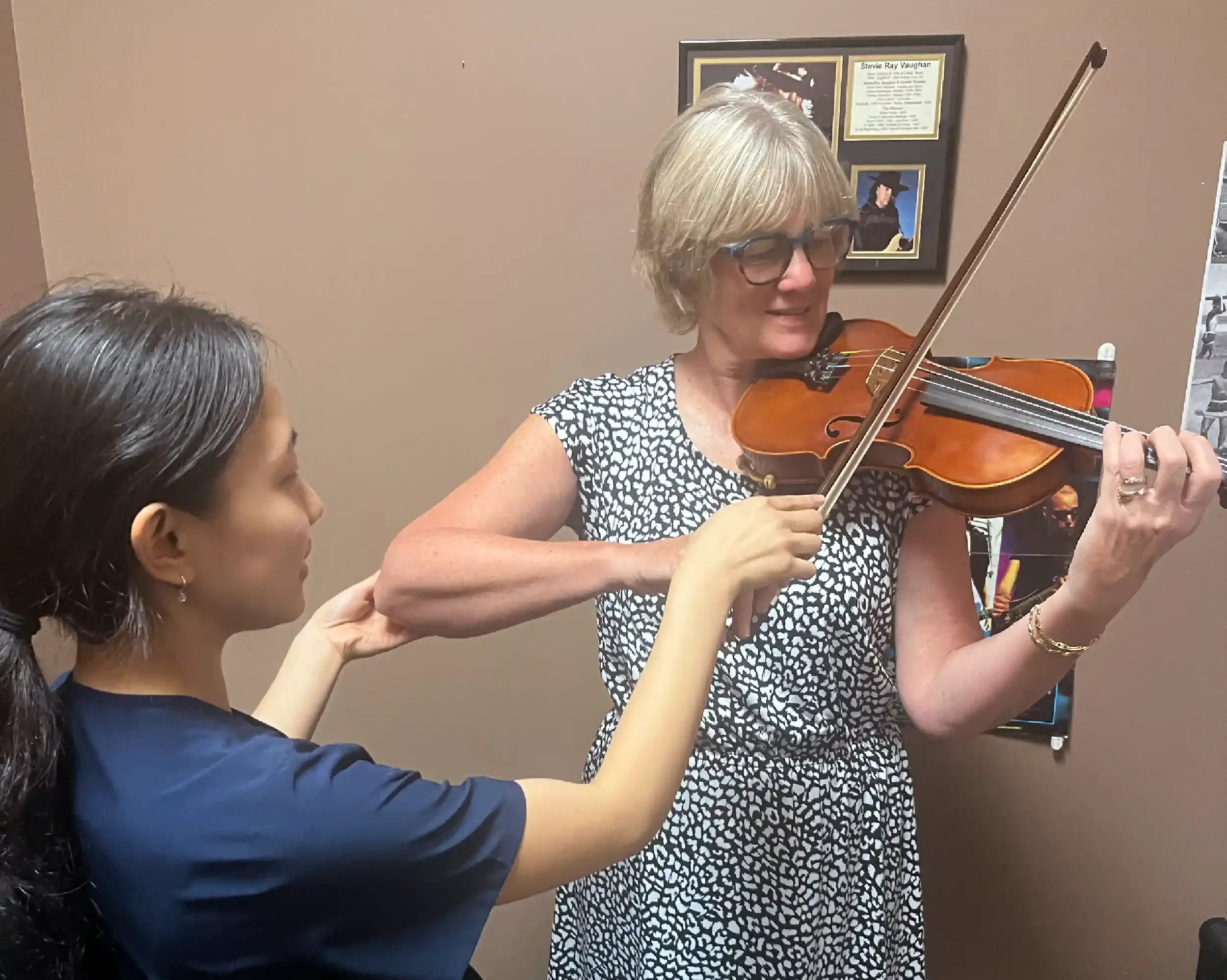 A woman with gray hair and glasses playing the violin while a young girl with dark hair in a ponytail assists or observes. They are indoors, with framed pictures on the wall behind them.