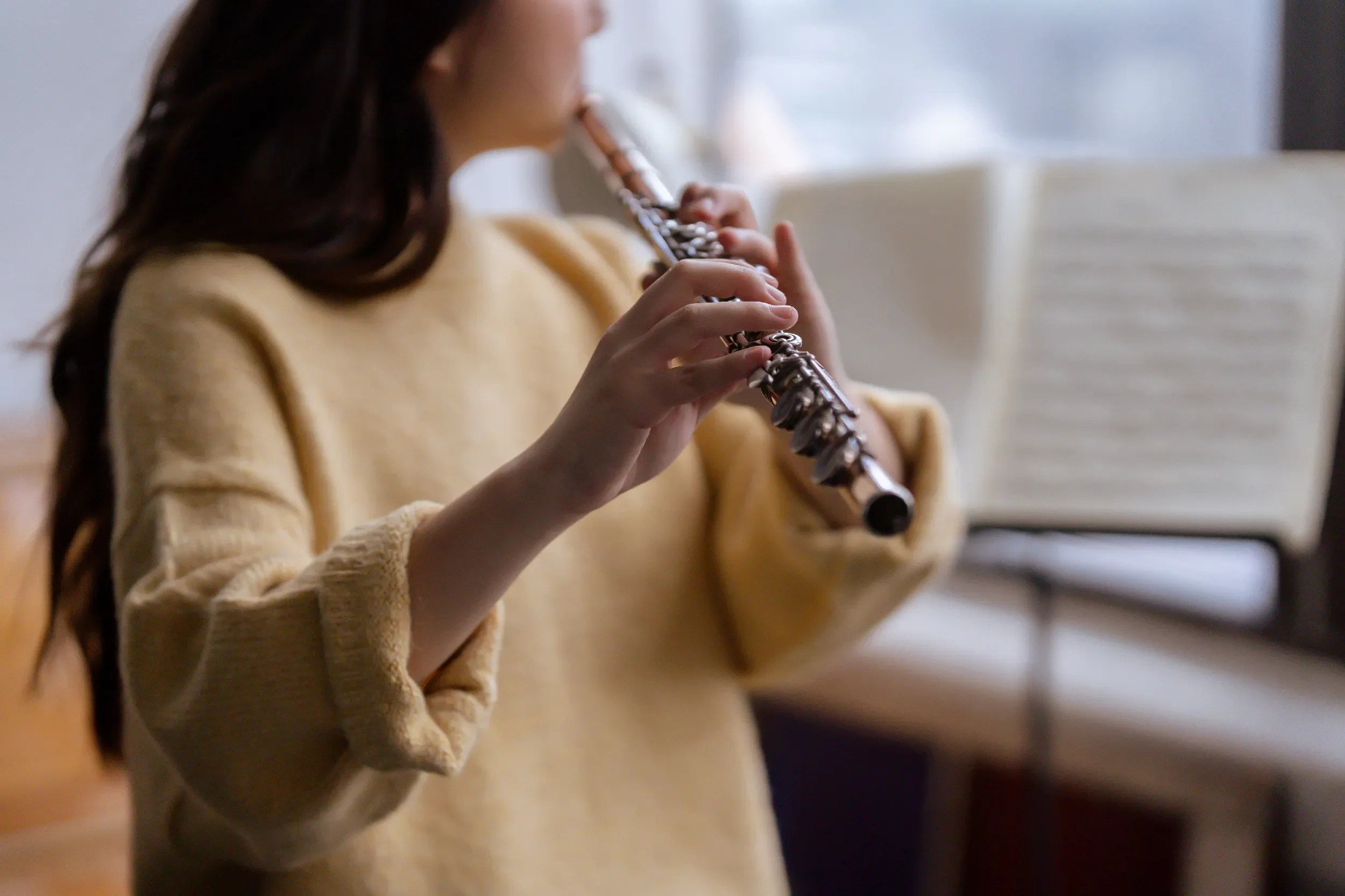 A woman with long dark hair wearing a beige sweater playing a silver flute.
