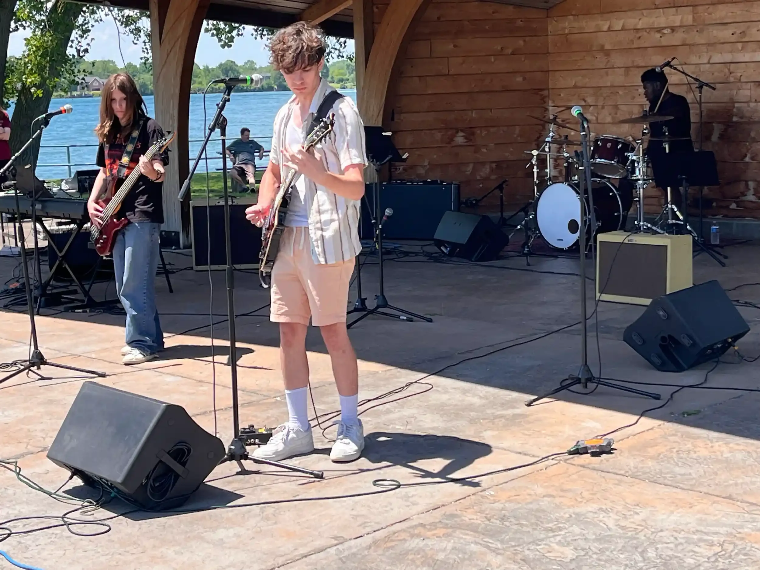 Young male musician with curly hair, wearing a striped shirt and shorts, playing an electric guitar on an outdoor stage near a lake, with a drummer and a female guitarist nearby, and a person sitting by the water in the background.