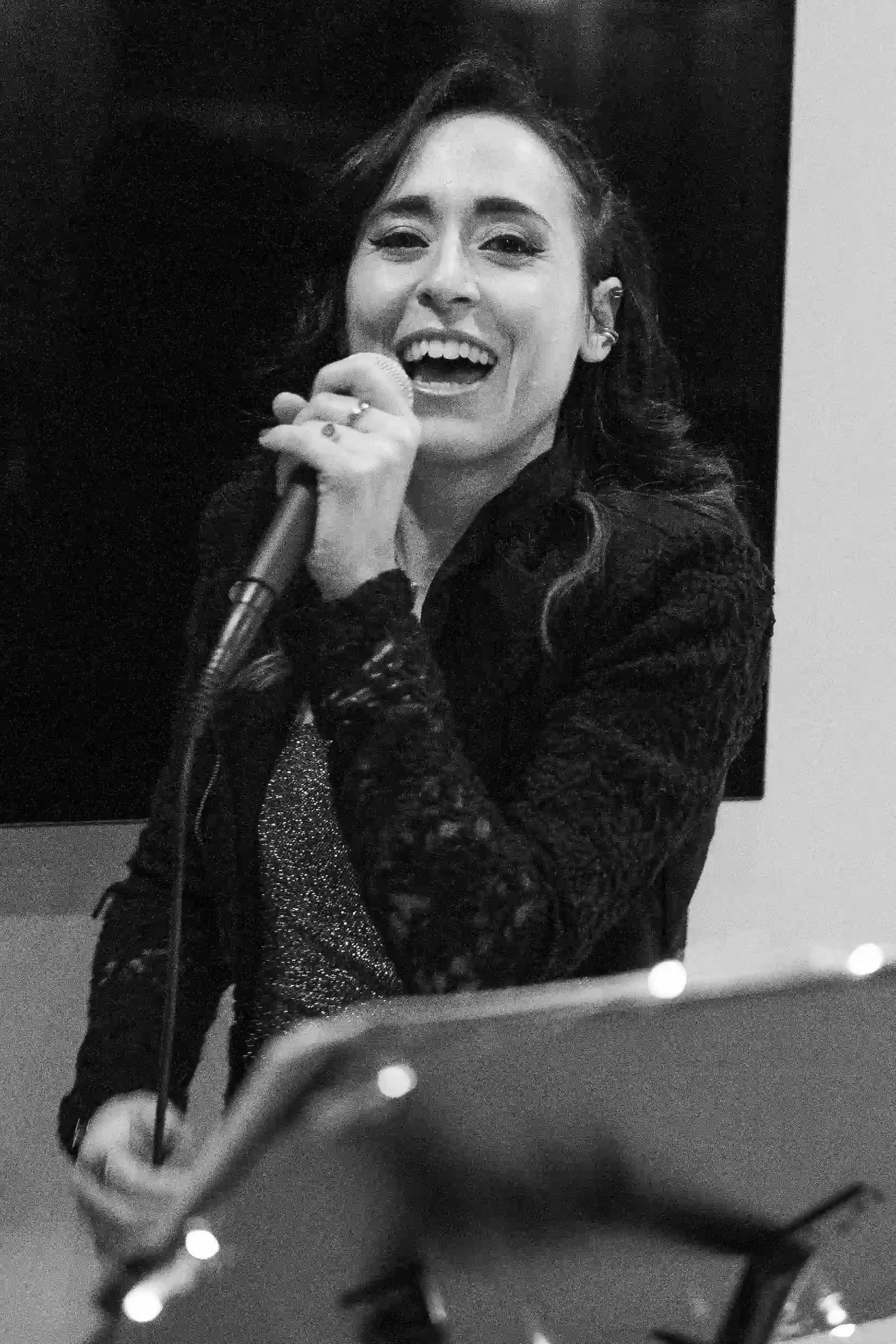 A woman with long hair holding a microphone, smiling and singing or speaking, dressed in a dark, patterned jacket, with earrings, photographed in black and white.