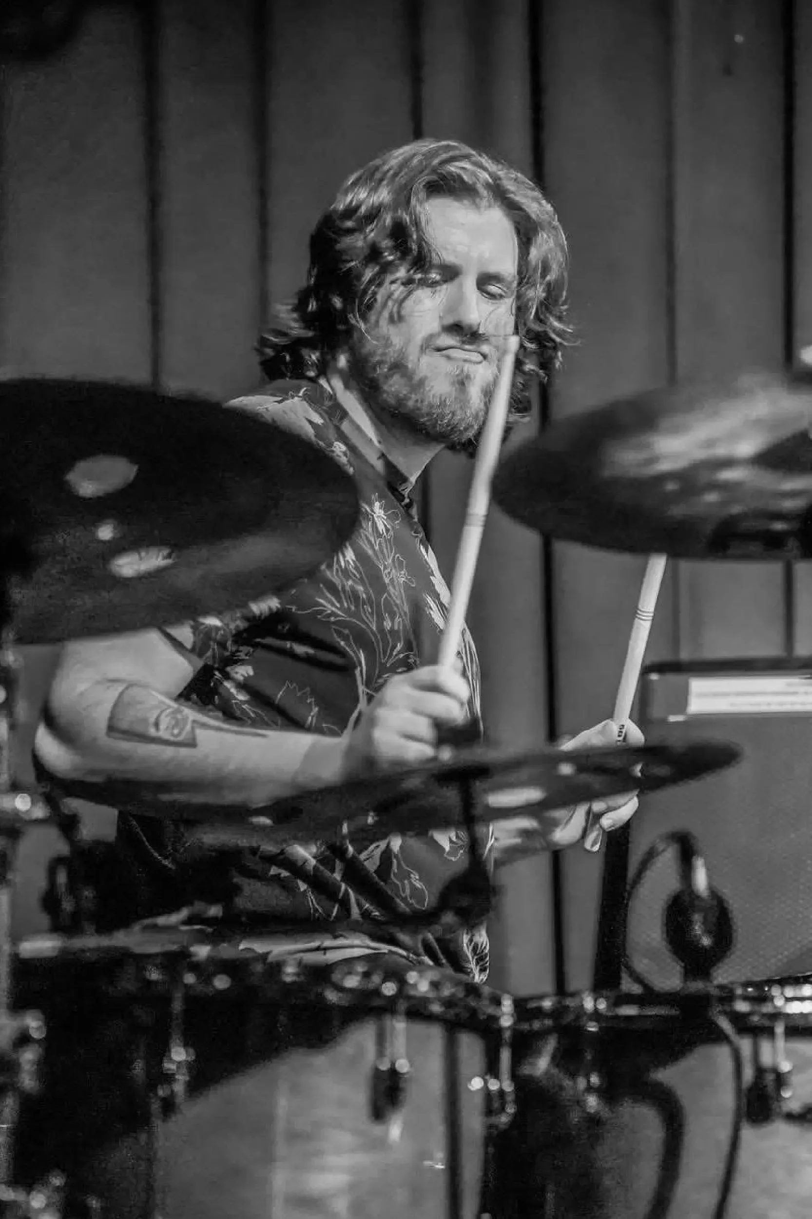 Black and white photo of a man with long wavy hair and a beard playing drums.