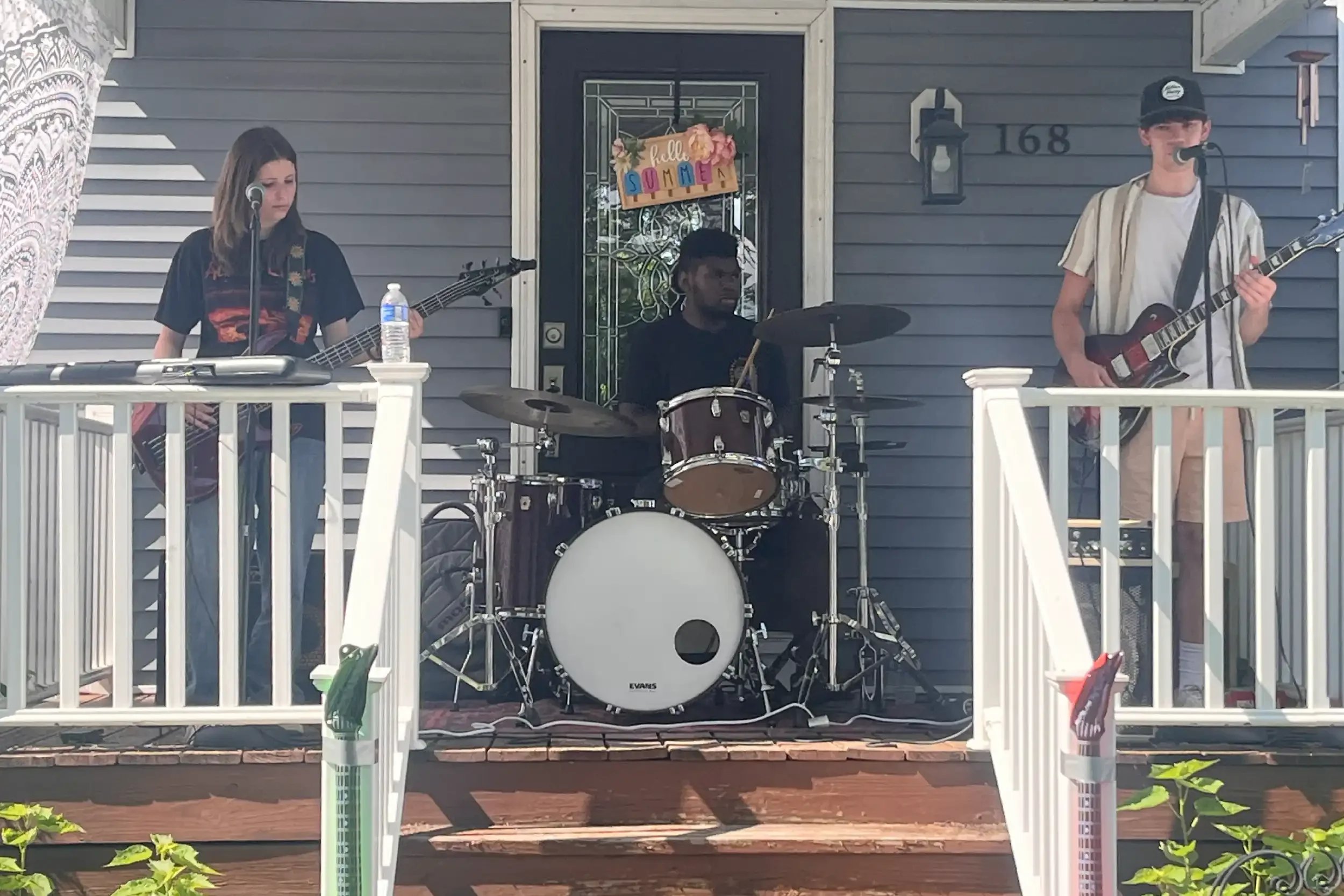 Three young musicians performing on a porch: a female guitarist, a male guitarist, and a male drummer. 