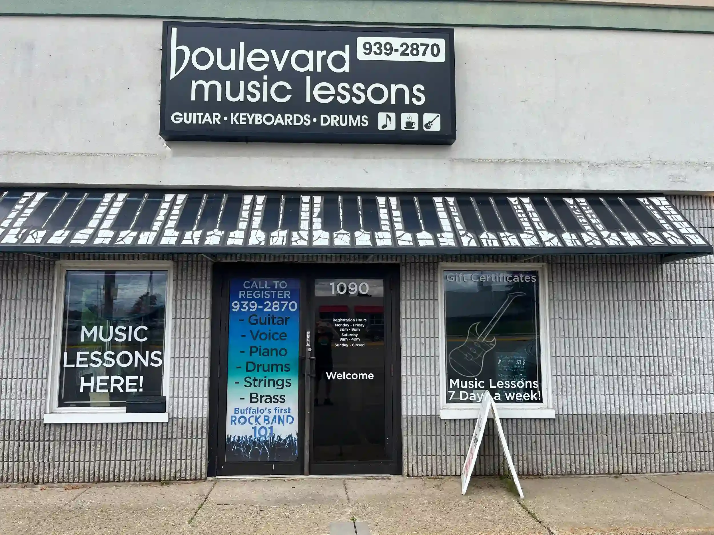 Storefront for music lessons with signs advertising guitar, keyboards, drums, voice, piano, strings, and brass lessons. The main sign displays the business name, phone number, and icons representing music instruments and sounds. The door has a sign w