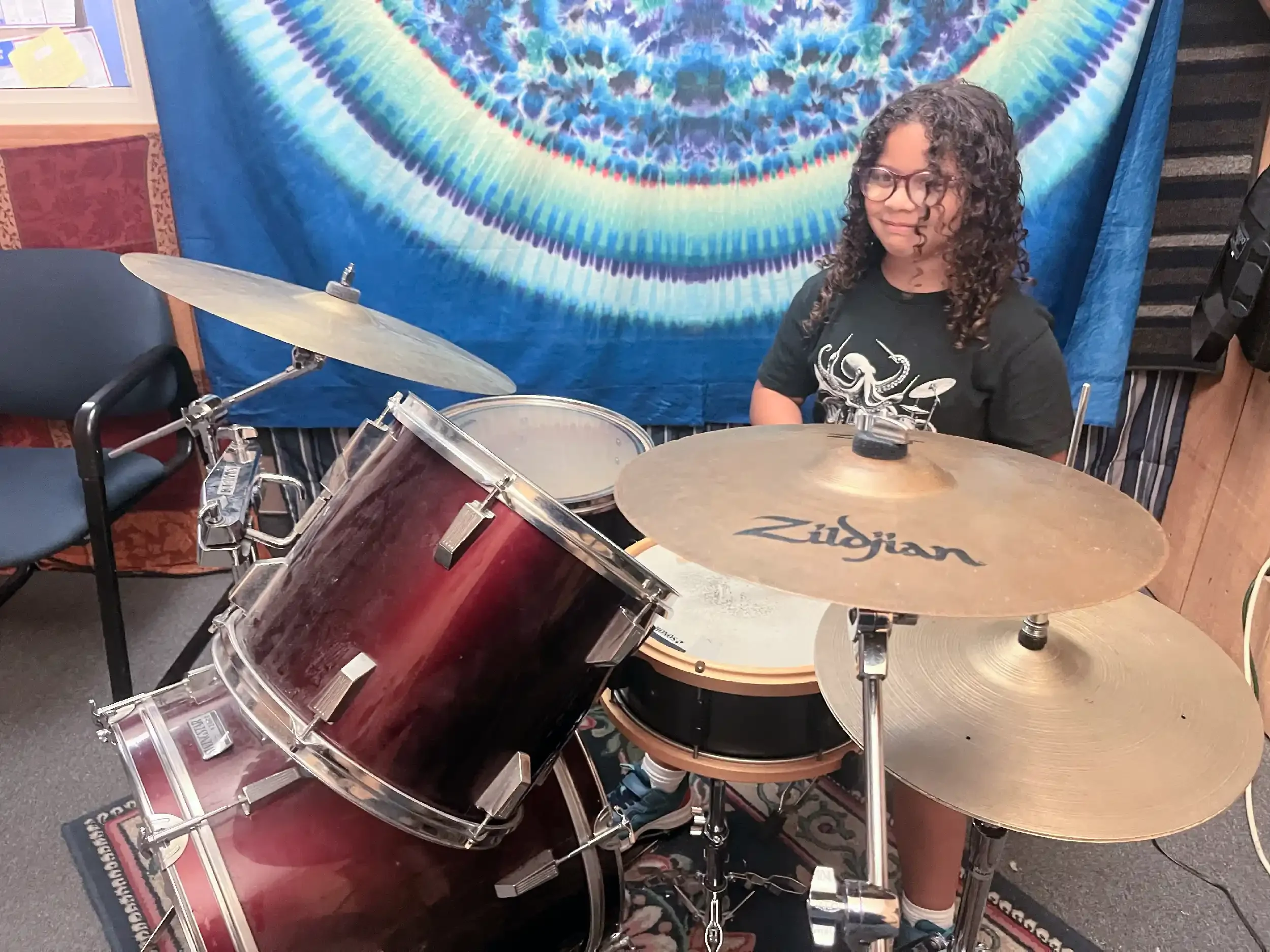 A young girl with curly hair and glasses, sitting behind a drum set, smiling. A colorful tie-dye tapestry hangs on the wall behind her.