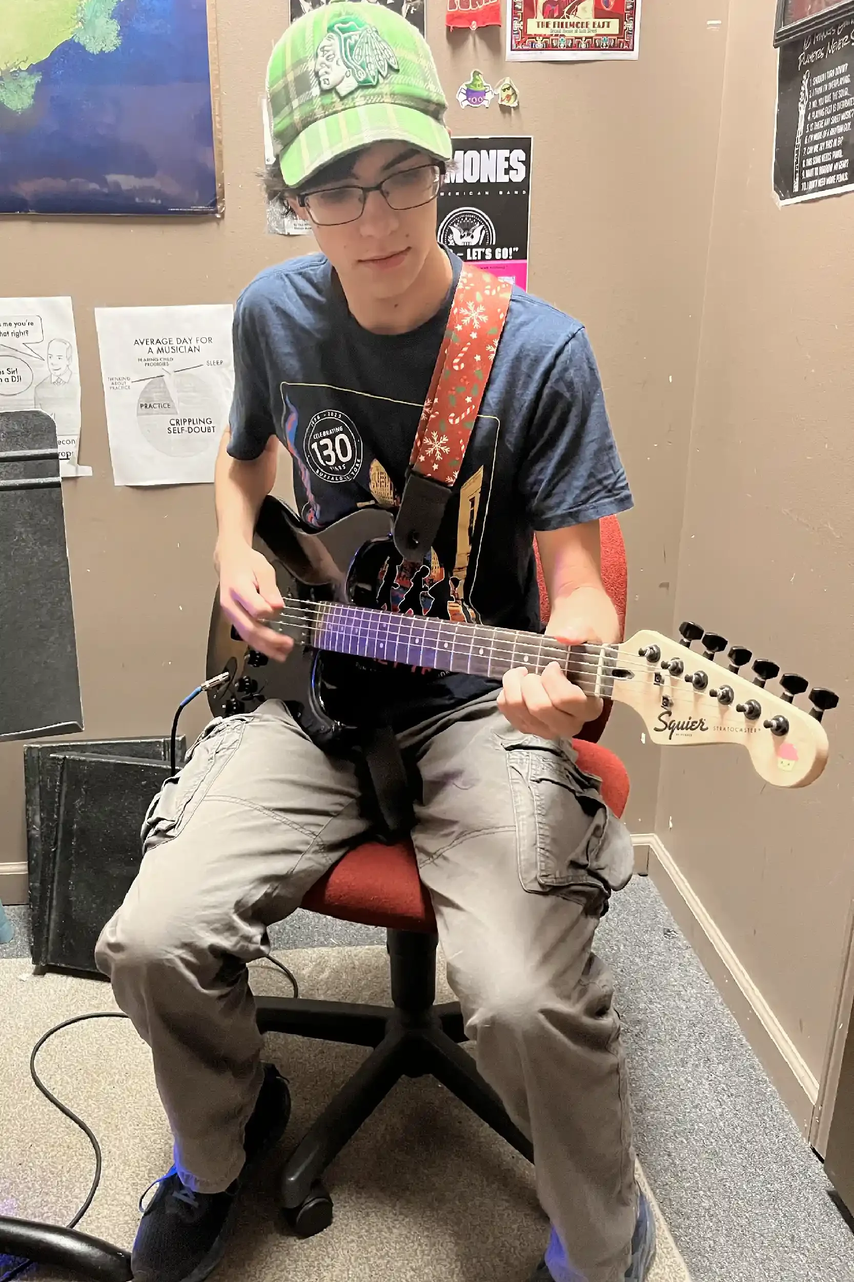 A young man wearing glasses and a green camouflage baseball cap sitting on a swivel chair. He is playing a black electric guitar, wearing a dark t-shirt and beige cargo pants. The setting appears to be an indoor room with musical posters and papers o