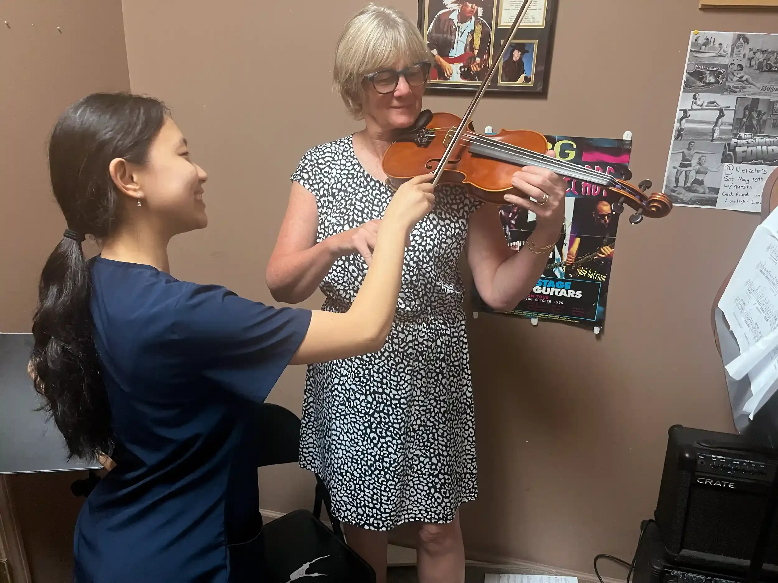 A woman playing the violin with a young girl assisting or learning from her, in a room decorated with music posters and photos on the wall.