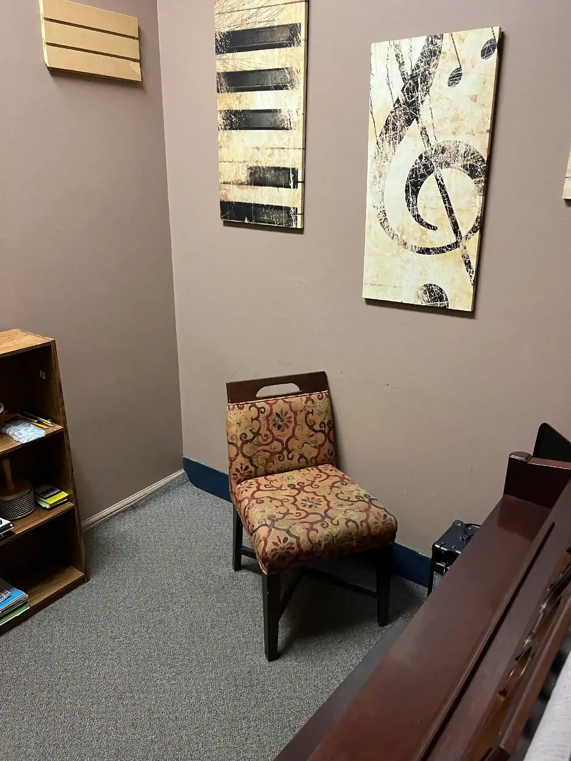 A small office corner with a patterned upholstered chair, a wooden bookshelf with books, and wall art featuring musical notes and abstract designs.