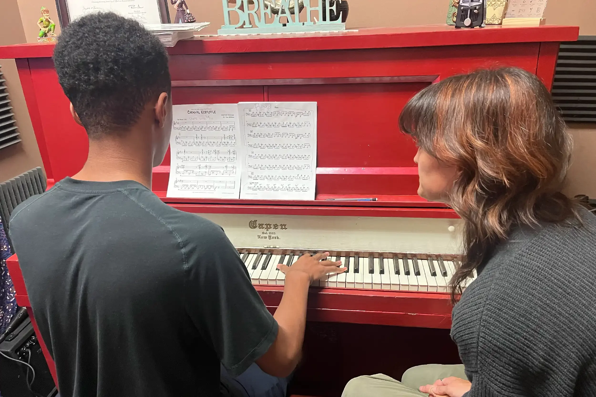 A young boy and a woman sit at a red piano, looking at sheet music titled 'Carnival Kerfuffle.' The boy's hand is on the piano keys, and they appear to be learning or practicing music together in a cozy room.
