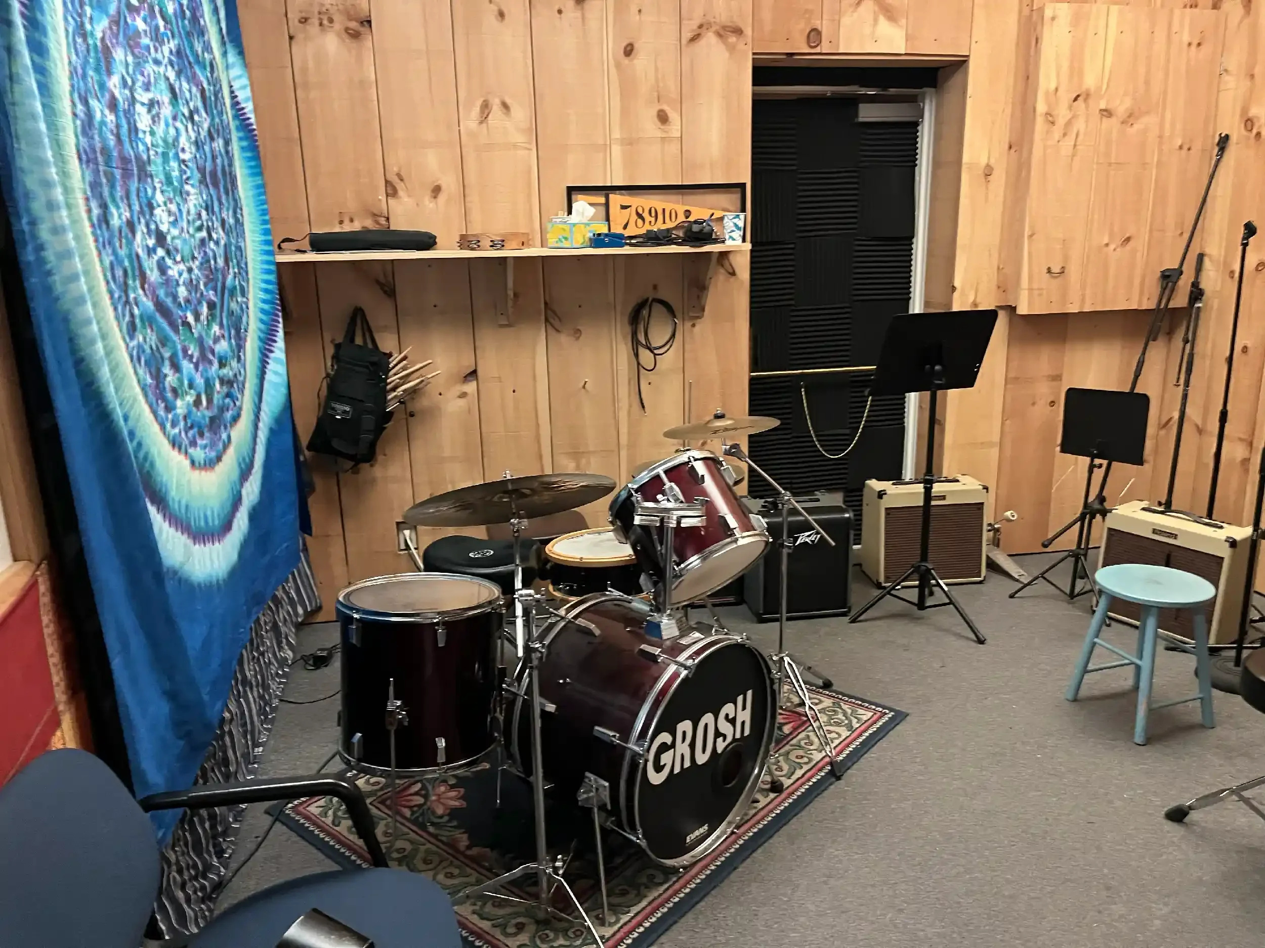 Music room with wooden wall paneling, blue tie-dye curtain on the left, and musical instruments including a drum set, guitar amplifier, and microphone stands. There is a small stool and a music stand, with various items and equipment on a shelf and t